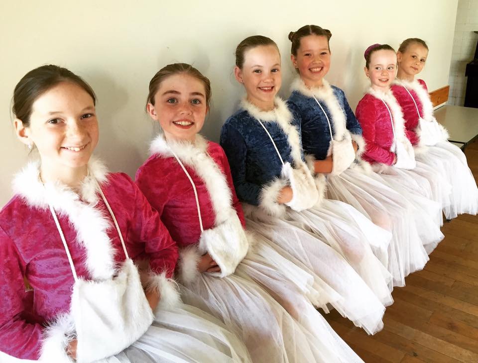 Six young girls dressed in holiday costumes, sitting in a row against a wall, smiling for the camera.