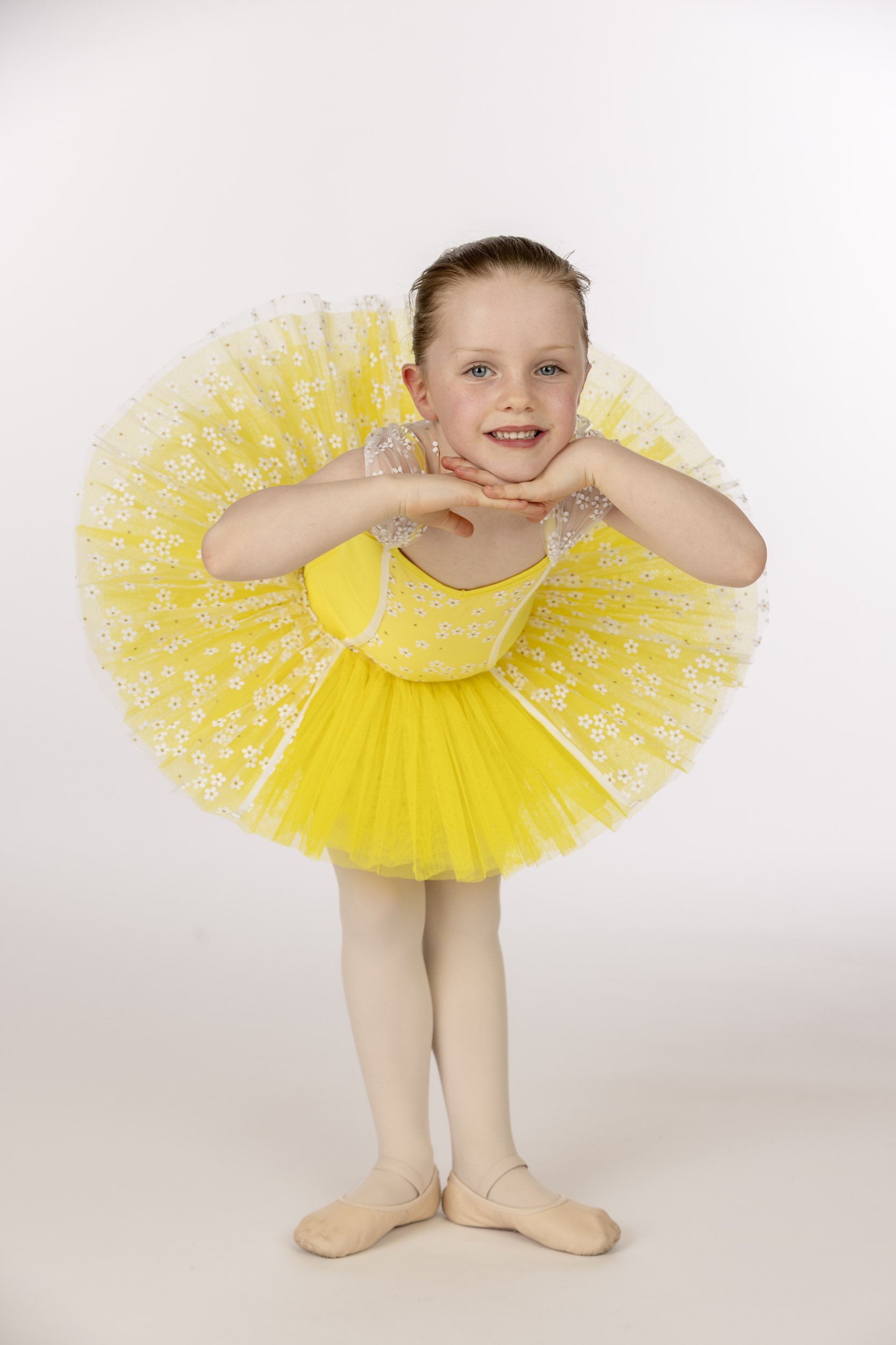 A young girl in a bright yellow ballet tutu and tights, posing with her hands under her chin, smiling, against a plain white background.