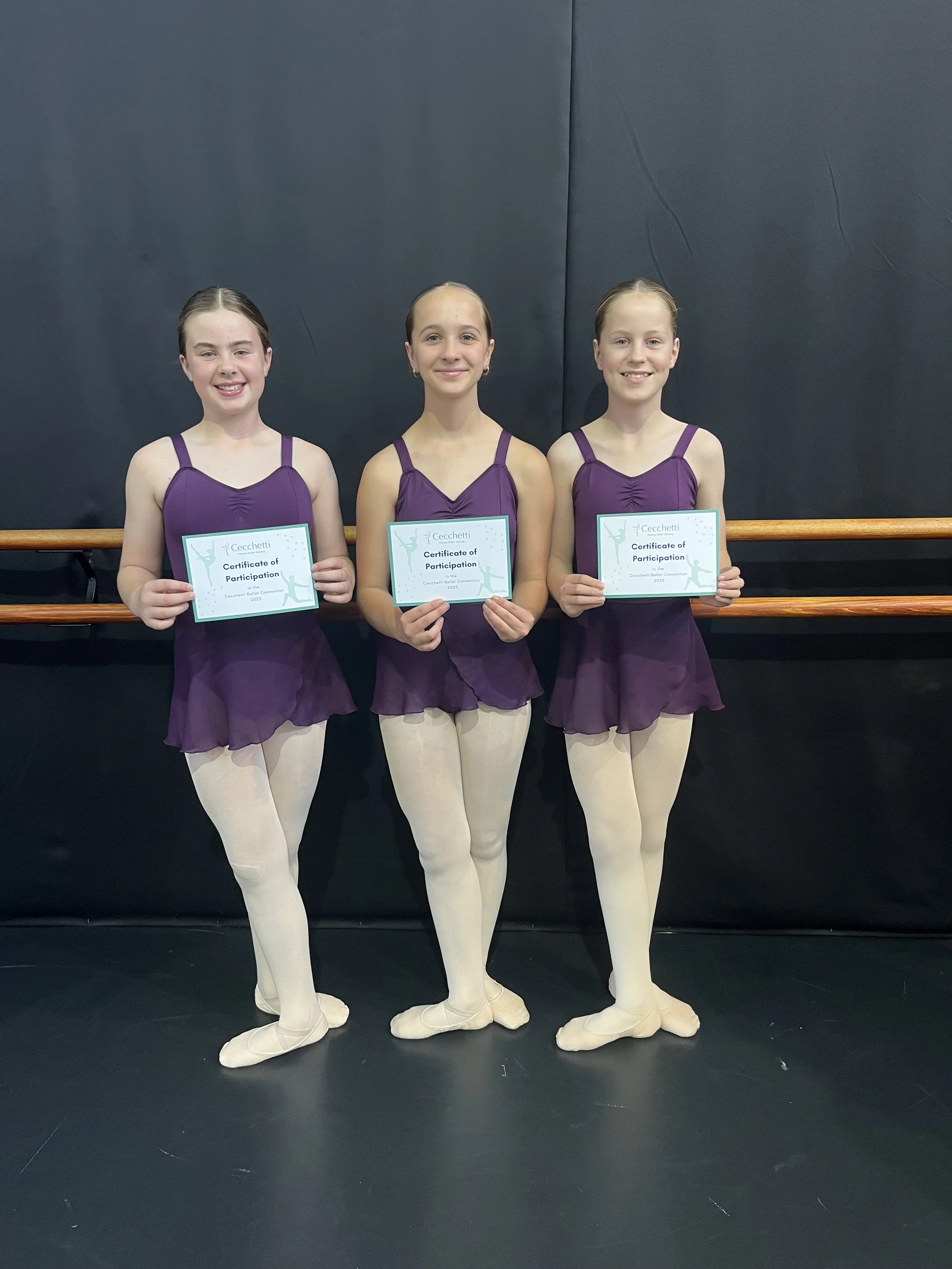 Three young girls in purple ballet costumes and white tights standing in front of a black wall holding certificates of participation from a ballet competition.