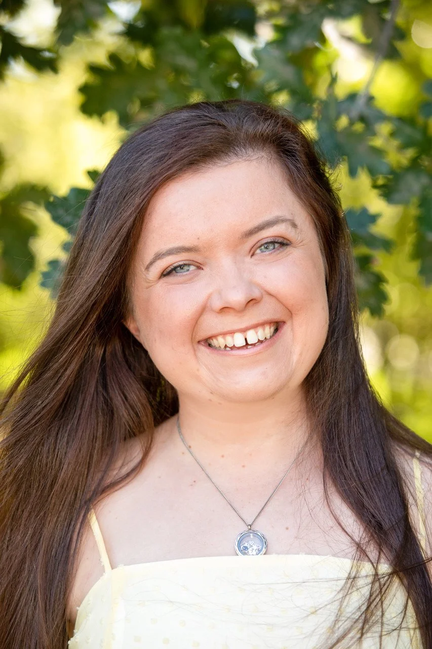 A woman with long brown hair and blue eyes smiling outdoors in front of green foliage.