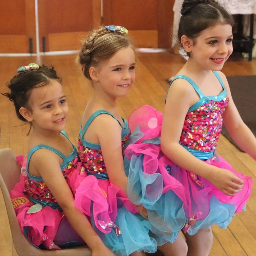 Three young girls dressed in colorful, sparkly pink and blue costumes sitting in a line, smiling, indoors with wooden floors.