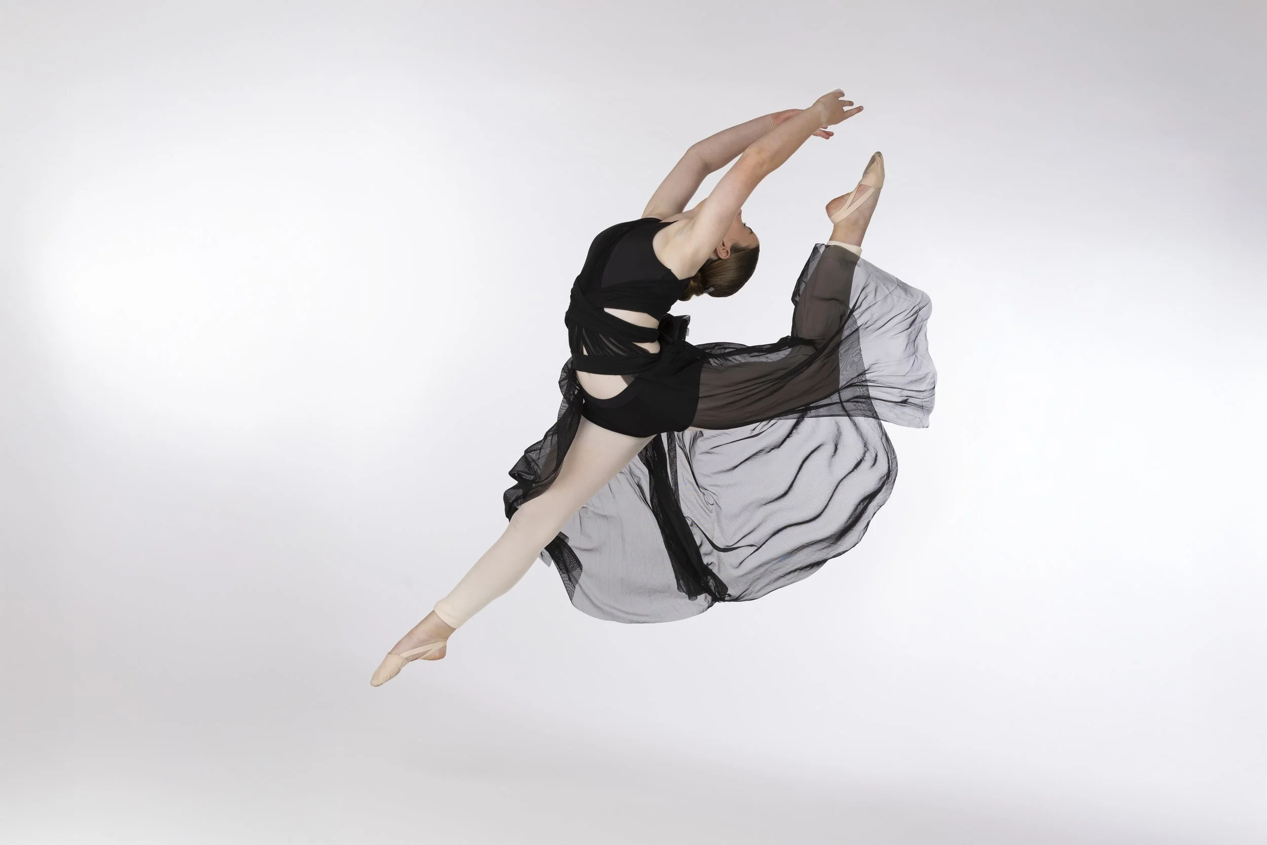 Ballet dancer performing a jump in a black and beige costume against a plain white background.