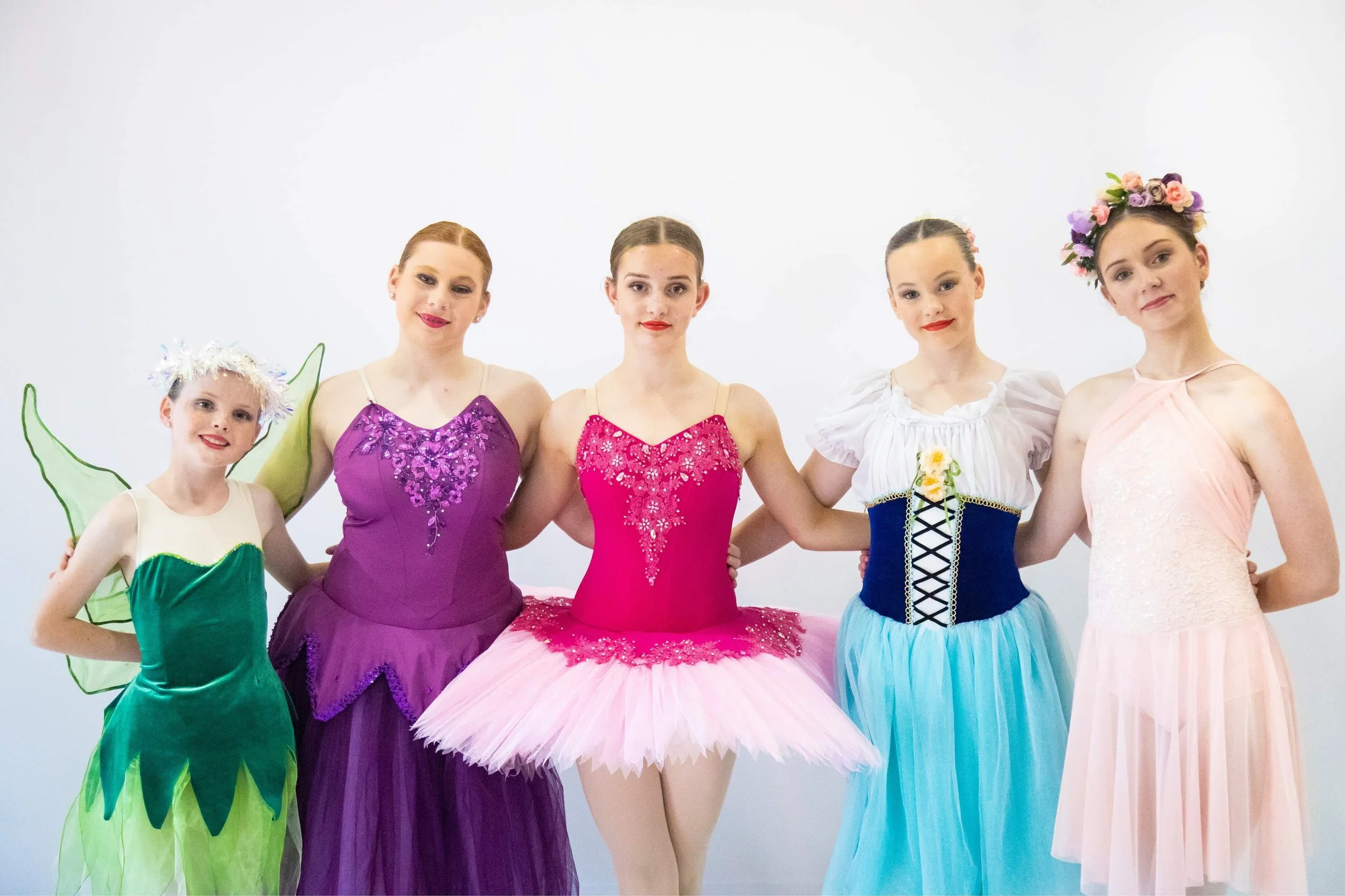Five young women and girls dressed in colorful ballet costumes stand together against a plain white background. The costumes include a green fairy, purple tutu, pink tutu, blue and white dress, and a peach-colored dress with a floral crown. They are 