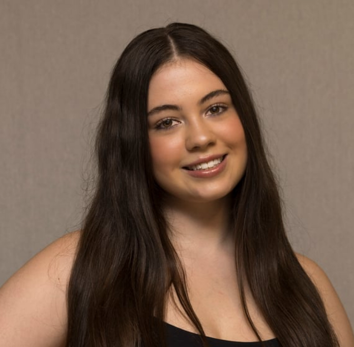 A young woman with long dark hair, smiling, wearing a black top, standing against a neutral background.