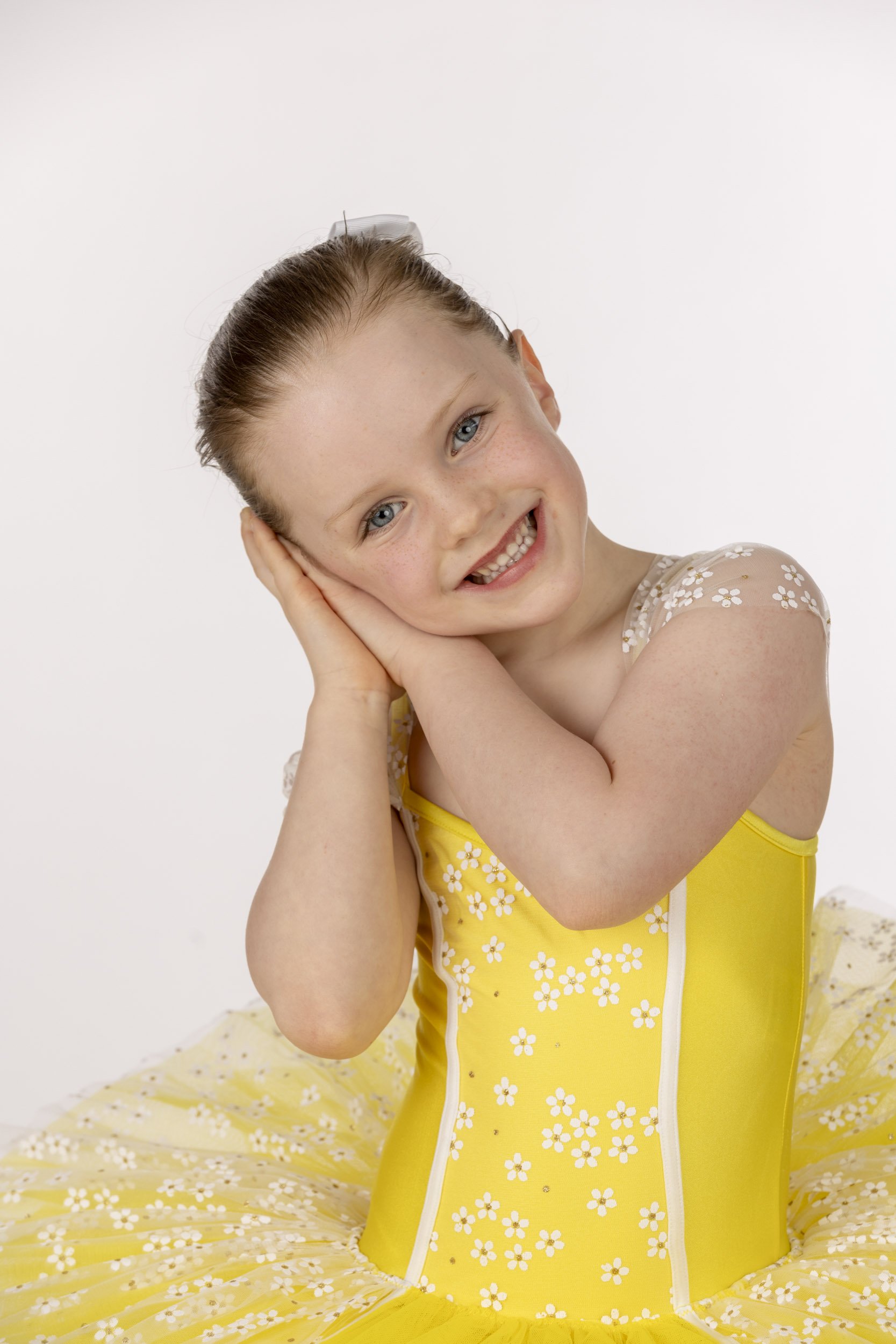 A smiling girl with blue eyes and red hair in a bun, wearing a yellow dress with white daisy patterns, sitting against a plain white background.