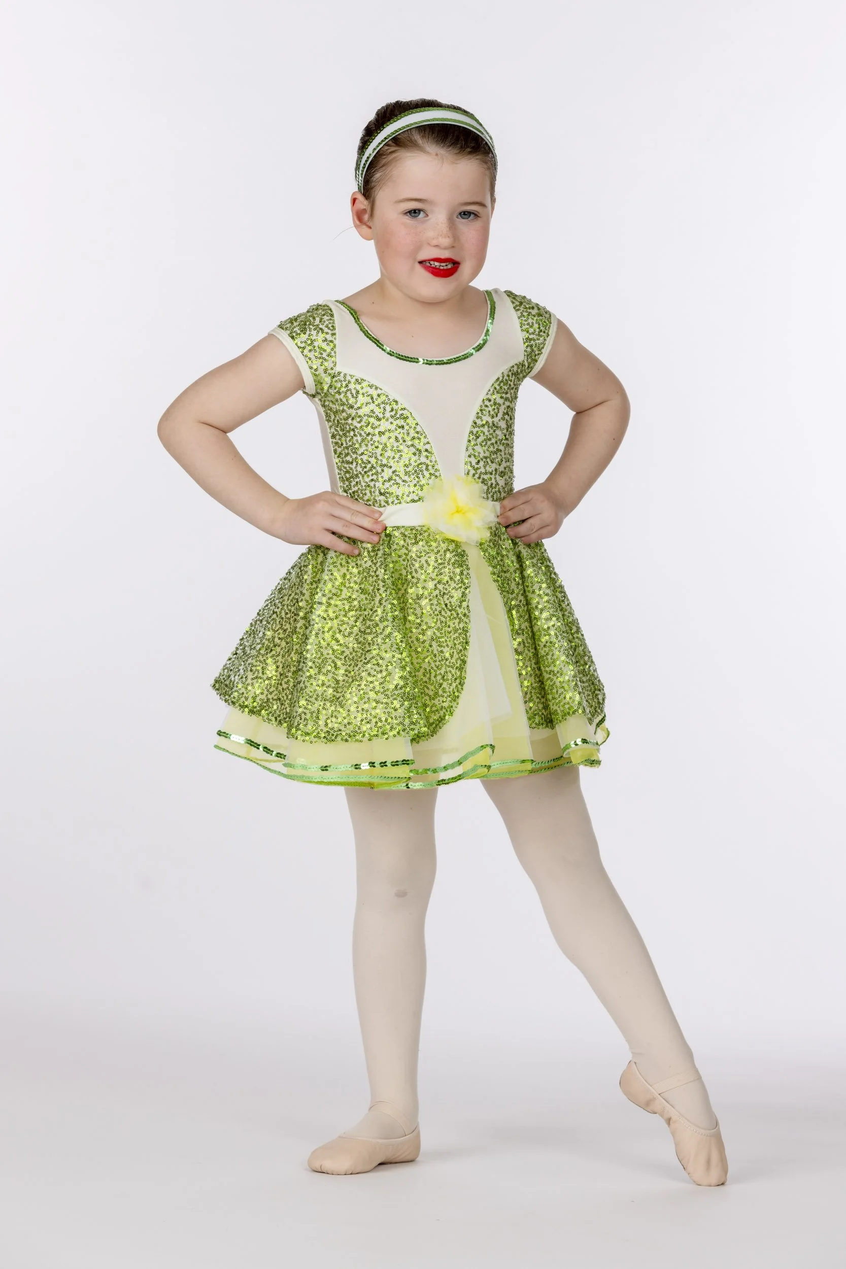 Young girl in a sparkly green and yellow dance costume with tights and ballet slippers, posing with hands on hips.