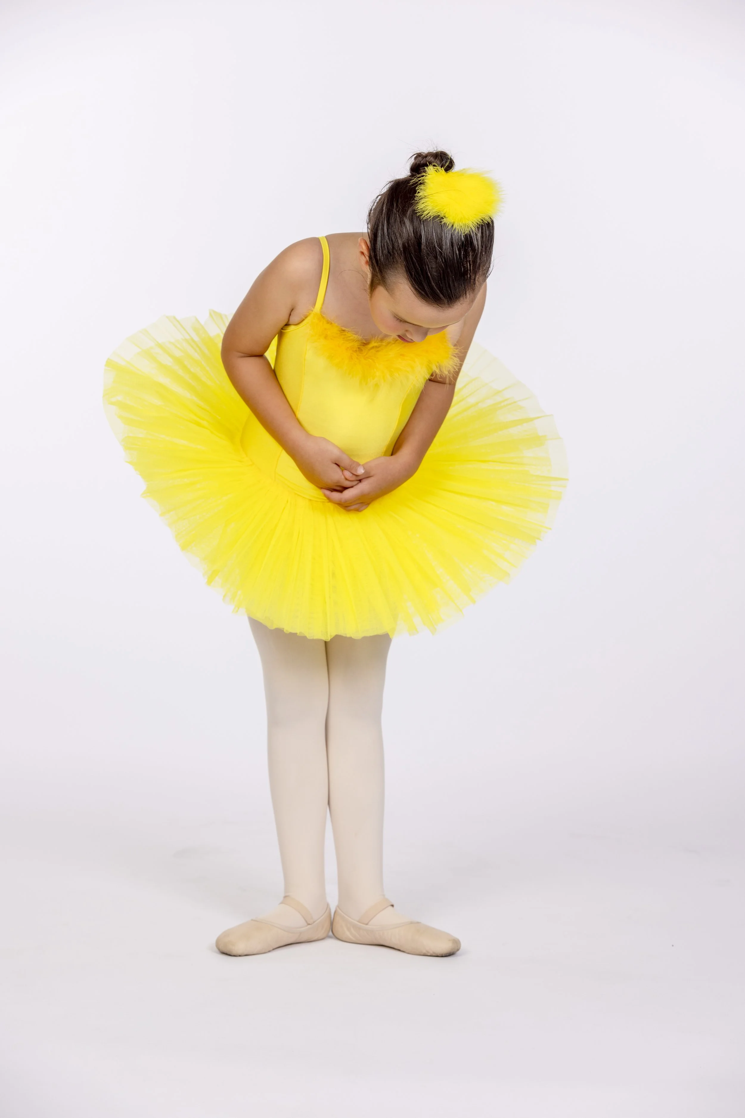 Young girl in a yellow ballet costume with a fluffy yellow hair accessory, standing with her head bowed and hands clasped in front, on a white background.