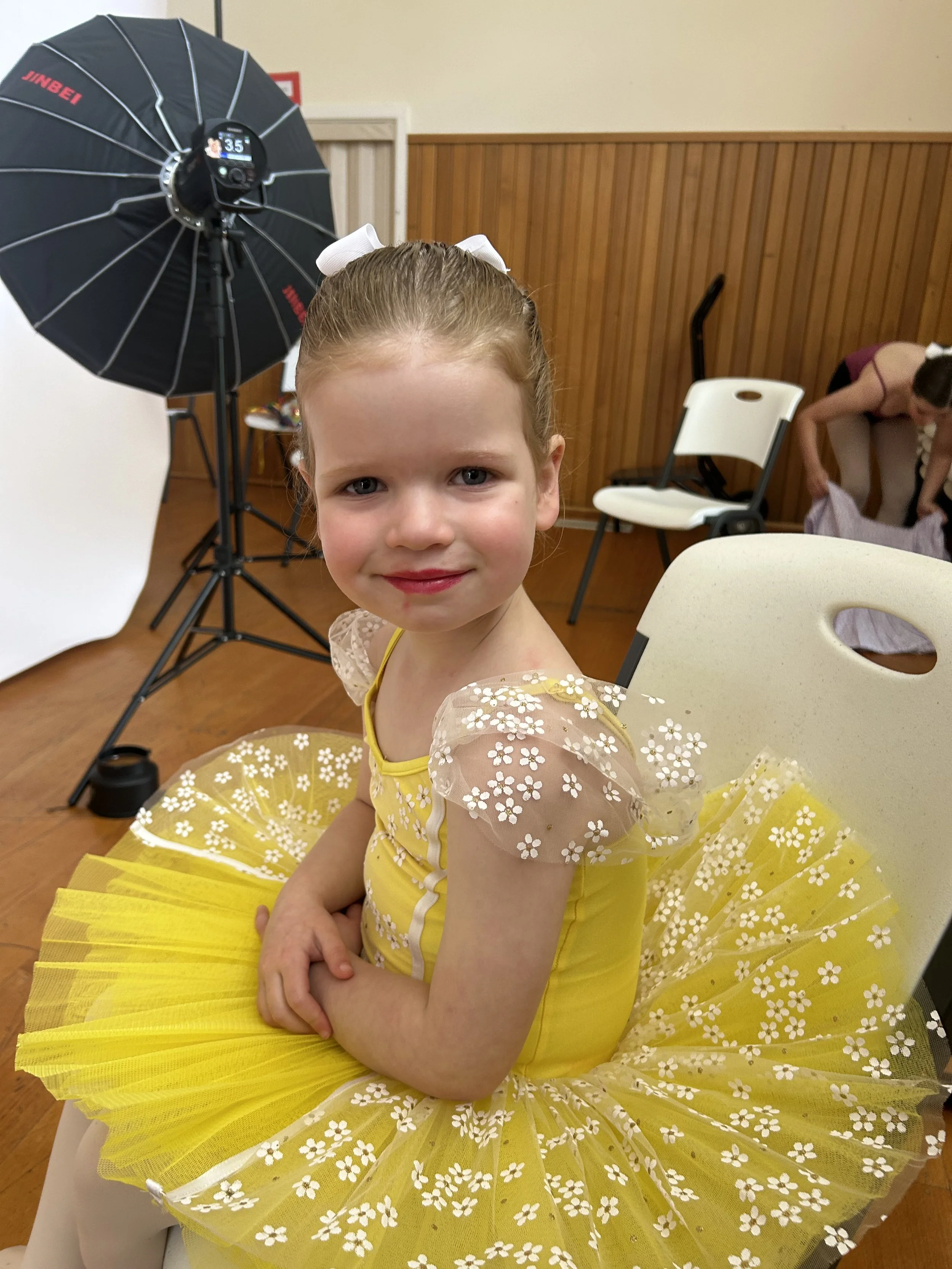 A young girl dressed as a ballerina in a yellow tutu with white floral patterns, sitting on a white chair in a dance studio or backstage area, with a photography lighting setup and another girl in the background