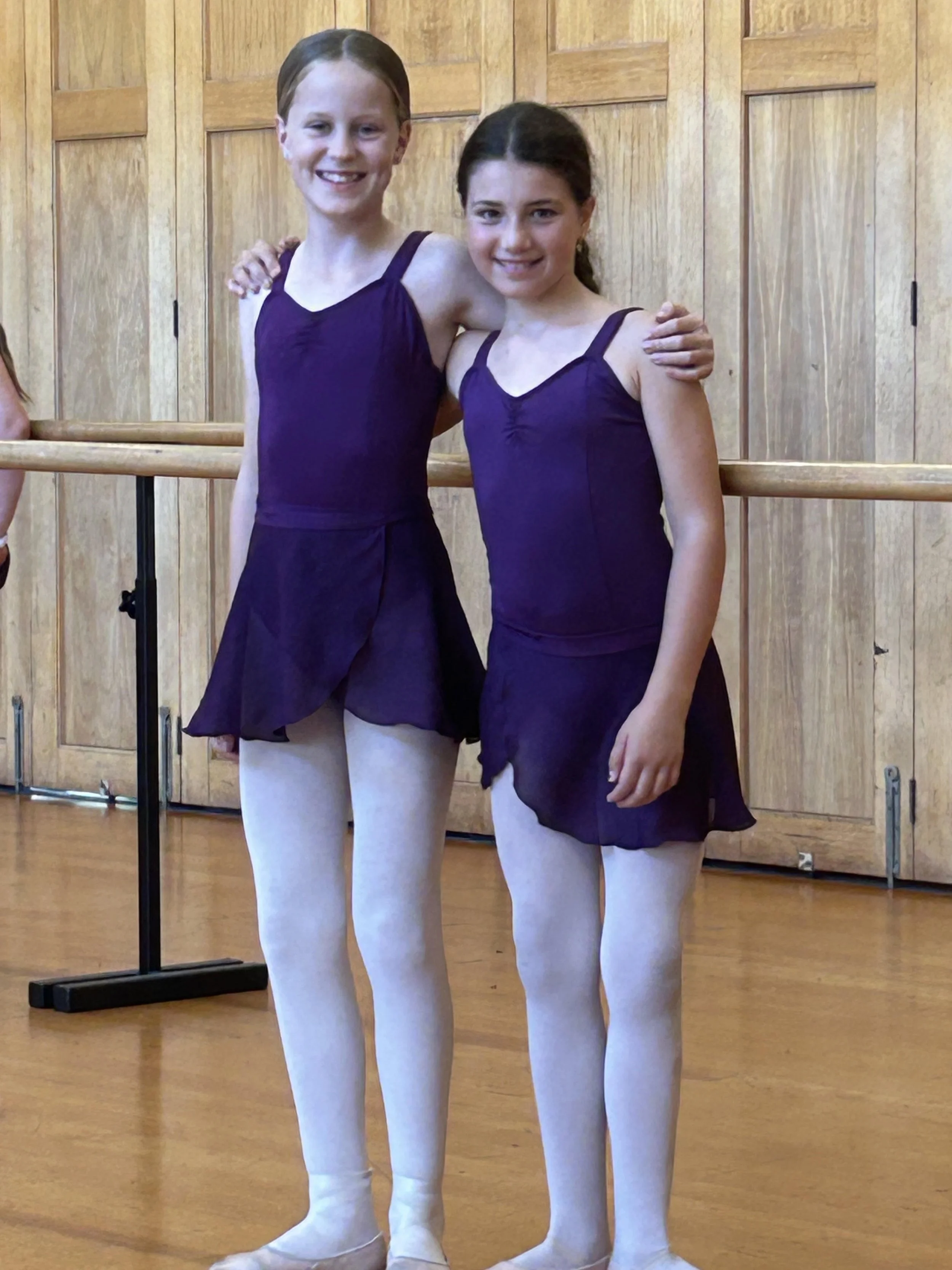 Two young girls in purple ballet outfits and white tights standing at a ballet barre in a dance studio, smiling at the camera.