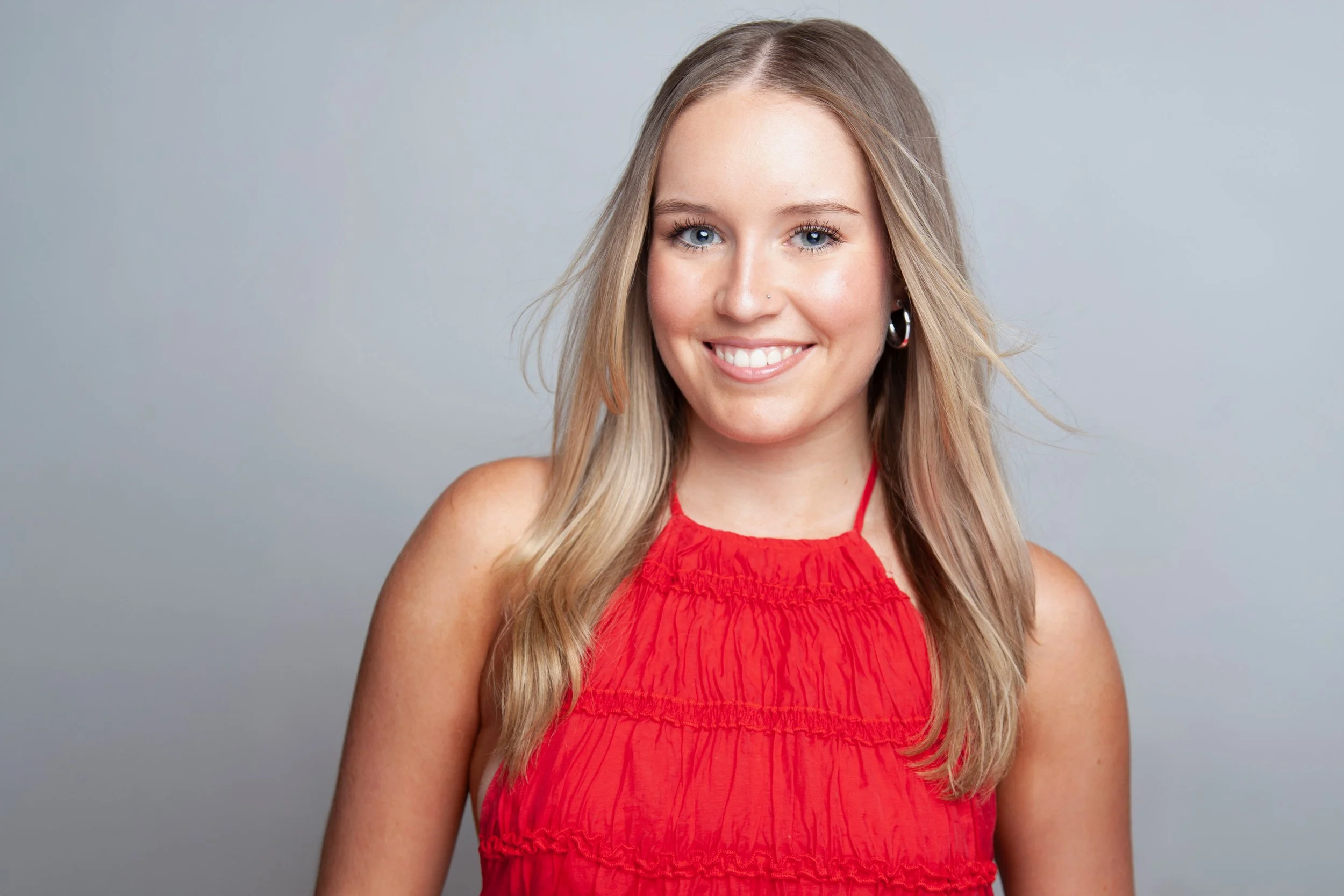 A young woman with long blond hair, blue eyes, freckles, and earrings, wearing a red sleeveless top, smiling at the camera against a plain light gray background.