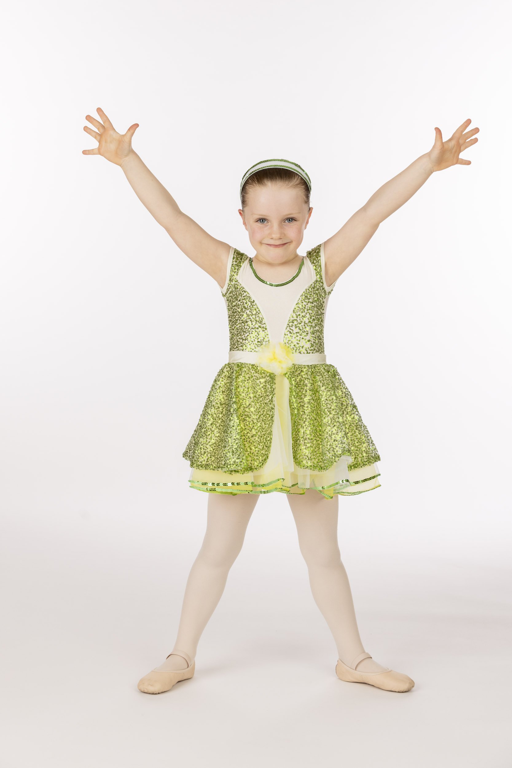 A young girl dressed in a sparkly green and yellow ballet costume with a matching headband, standing with her arms raised in a ballet pose on a plain white background.