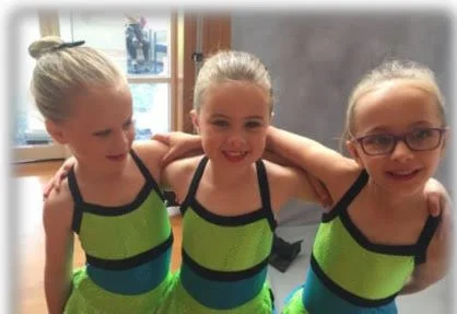 Three young girls in matching green and black leotards standing together with arms around each other, smiling in a dance studio.