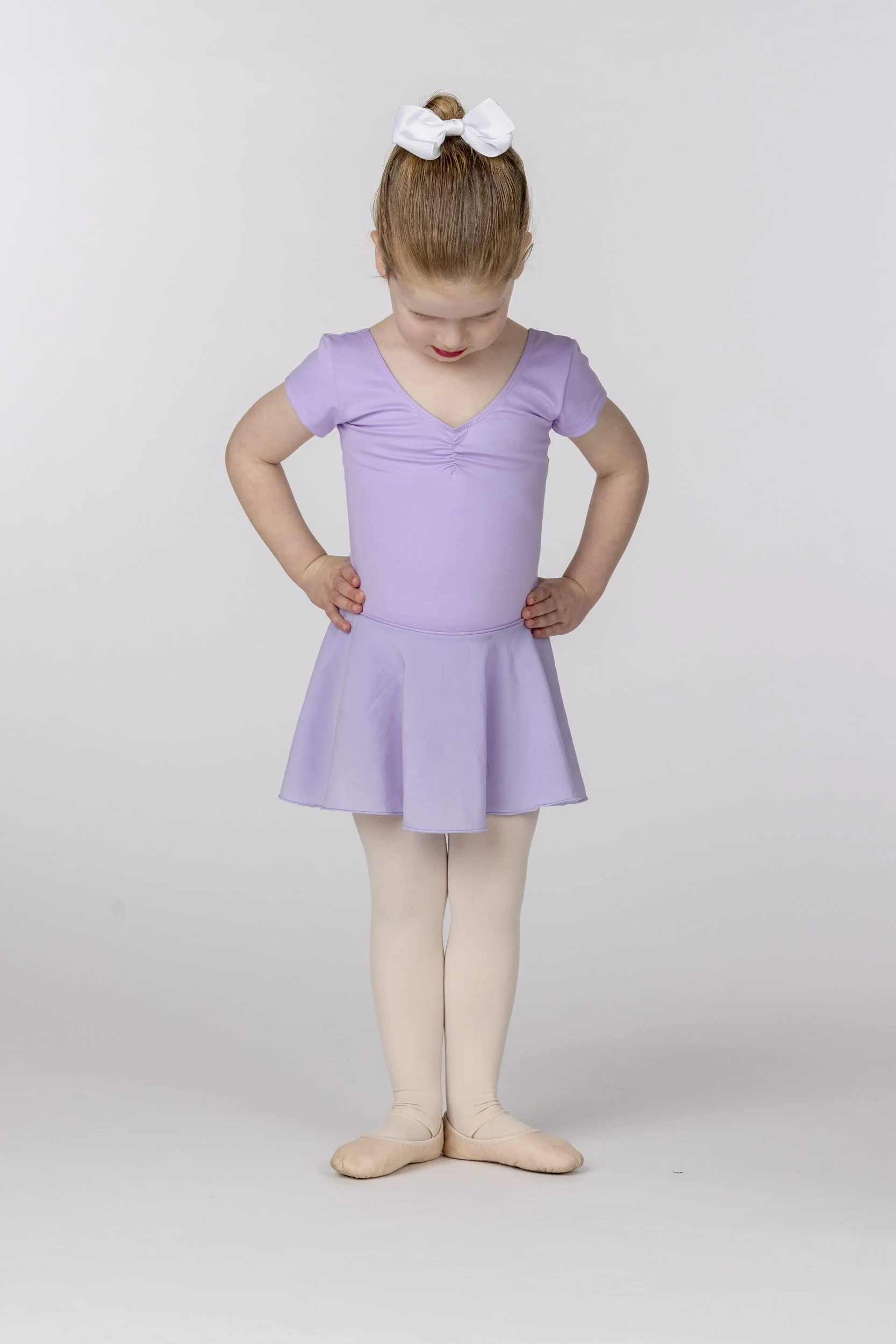 Young girl in a lavender ballet outfit, white tights, and beige ballet slippers, standing with her hands on her hips and looking down, against a plain white background, with her hair tied up with a white bow.