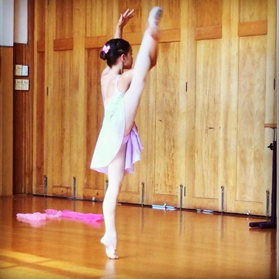 A young girl in a ballet pose with one leg extended high and arms raised, in a dance studio with wooden walls.