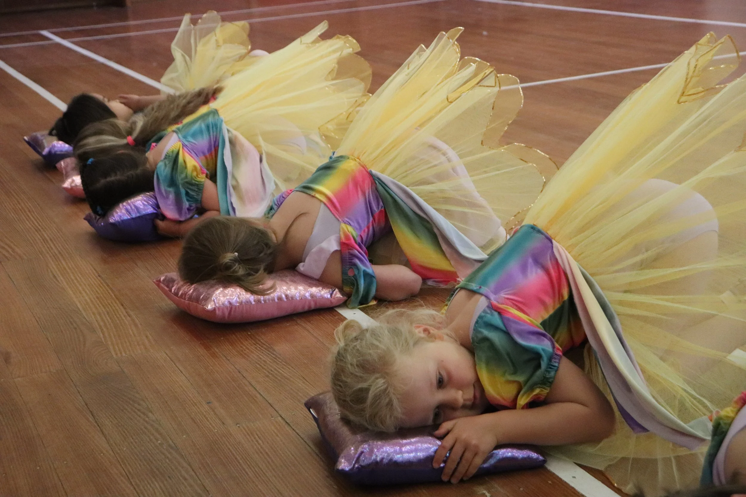 Young girls dressed as princesses in rainbow-colored tutus, lying on pink pillowcases on a gymnasium floor, resting with their eyes closed.