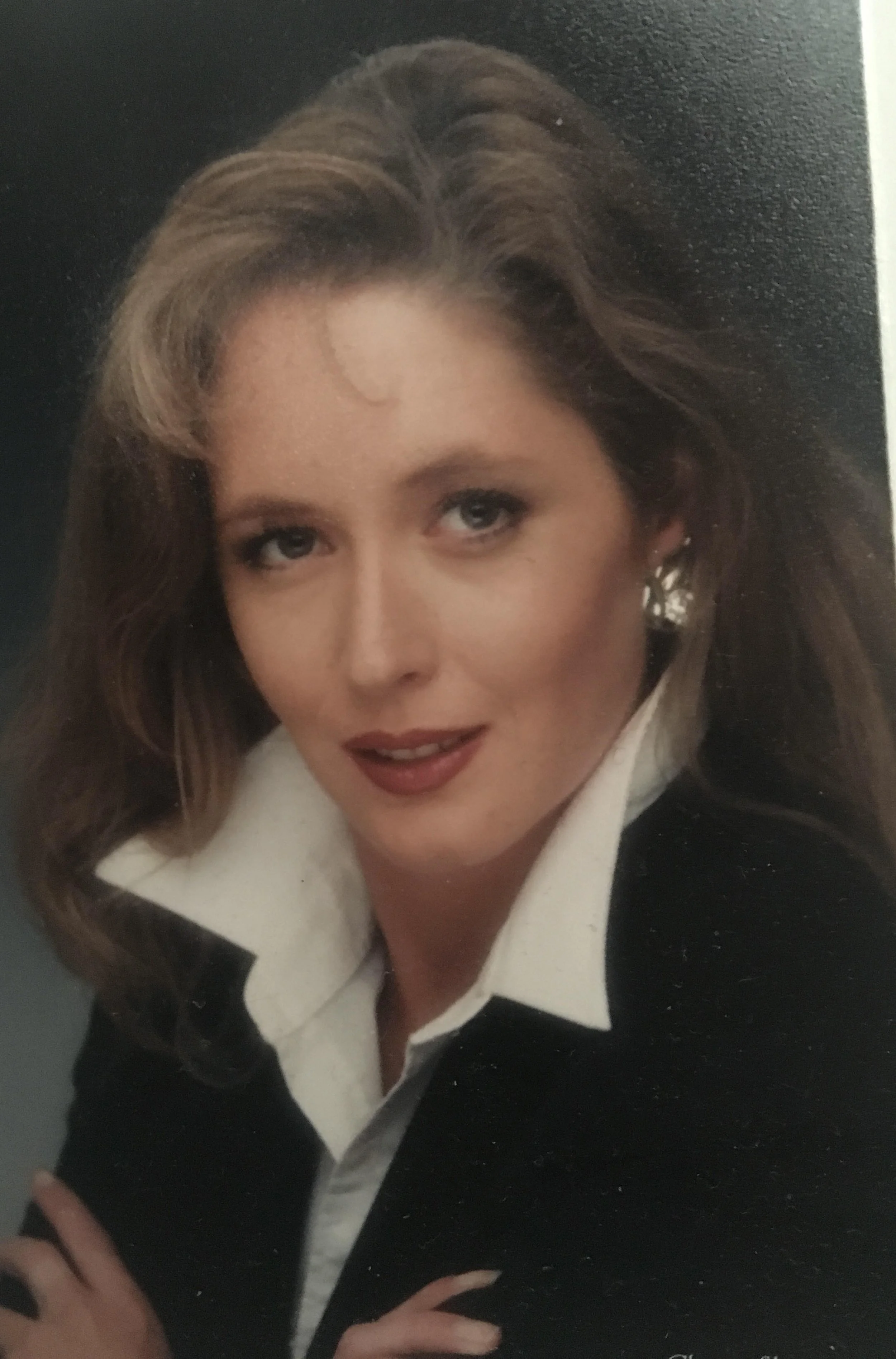 A woman with shoulder-length light brown hair, wearing a white collared shirt and black blazer, looking directly at the camera against a dark background.