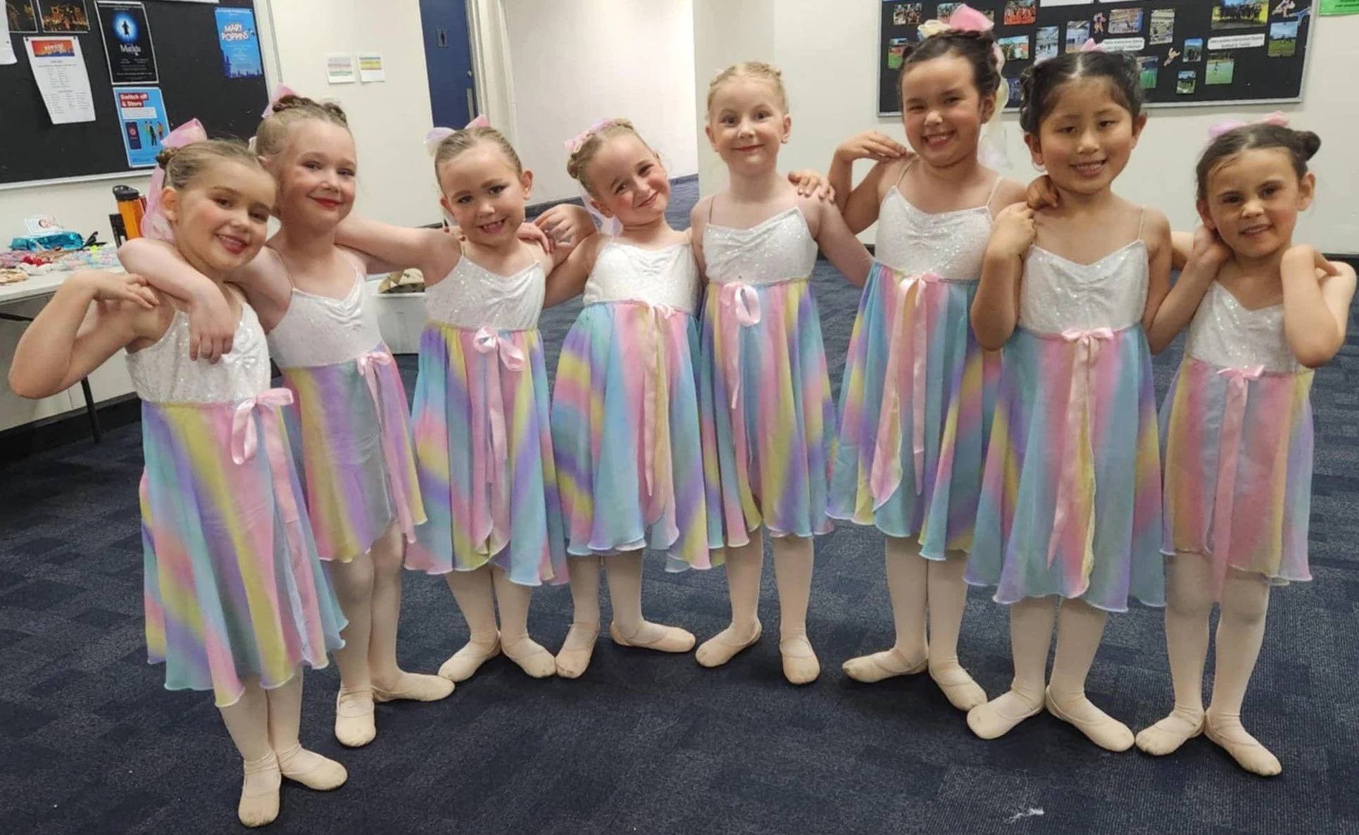Group of nine young girls in pastel rainbow ballet dresses and tights, posing together with arms around each other, smiling in a dance studio or backstage area.