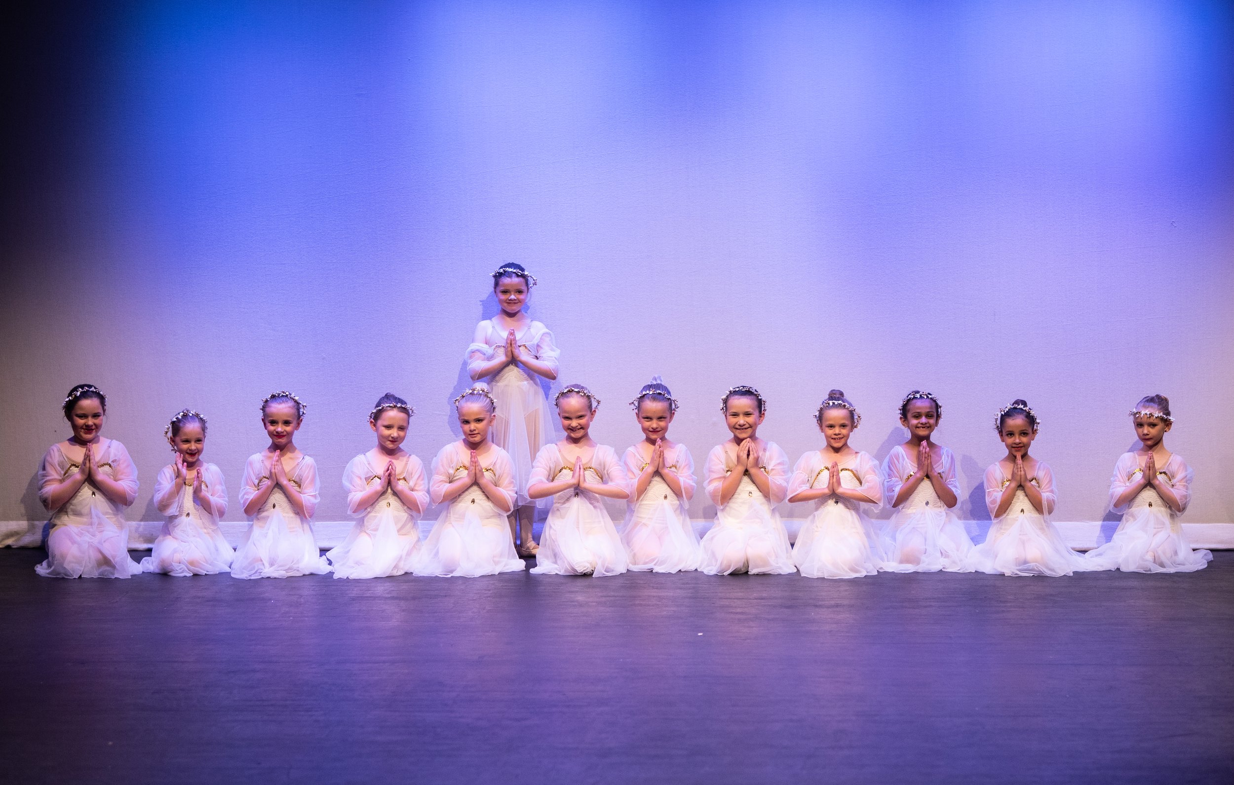 A group of young girls dressed in white ballet costumes with floral headbands, arranged in a line on a stage, some kneeling and one standing in the middle, all with hands in prayer pose, during a dance recital.