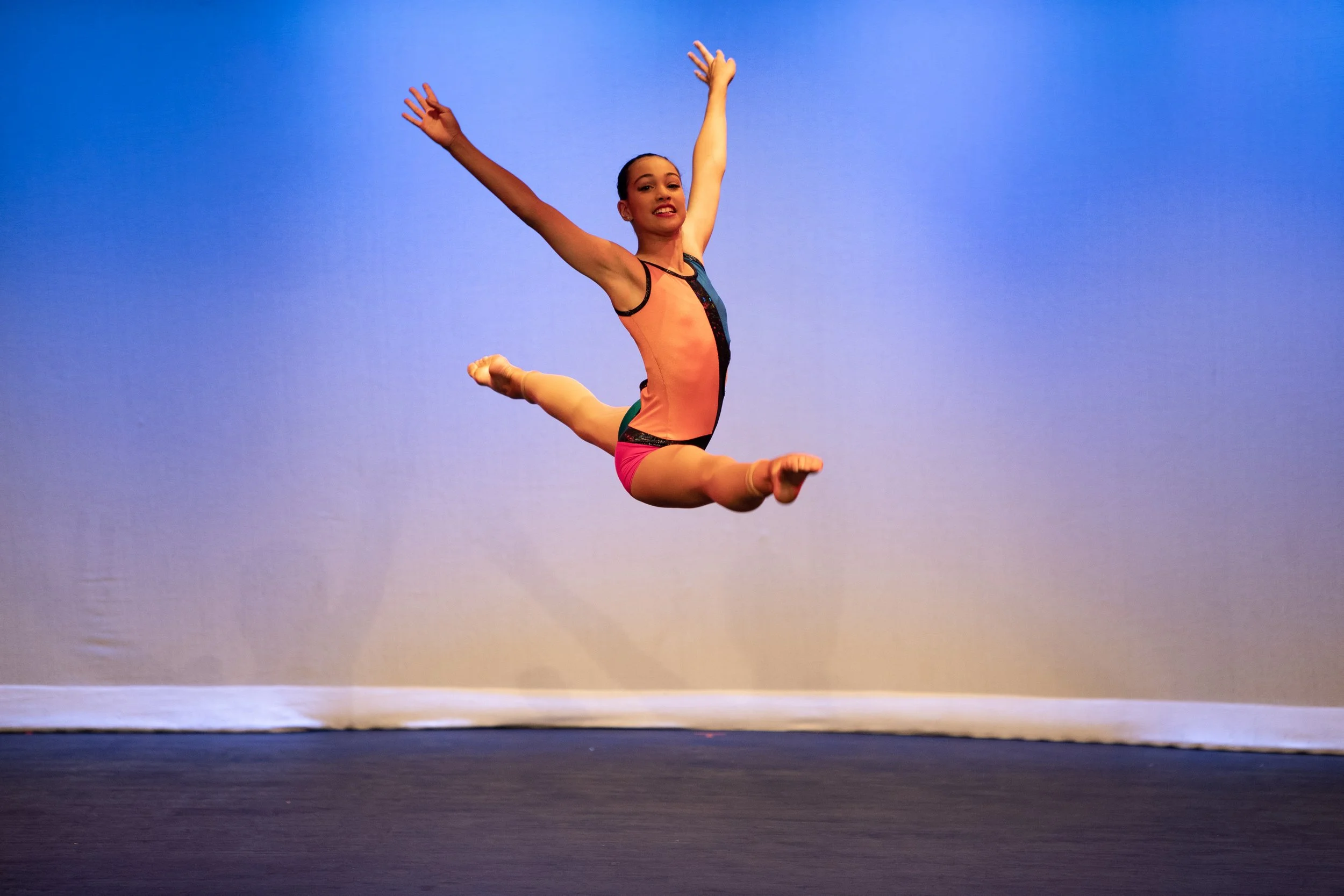 A young female gymnast mid-air during a leap on stage with a blue gradient background.