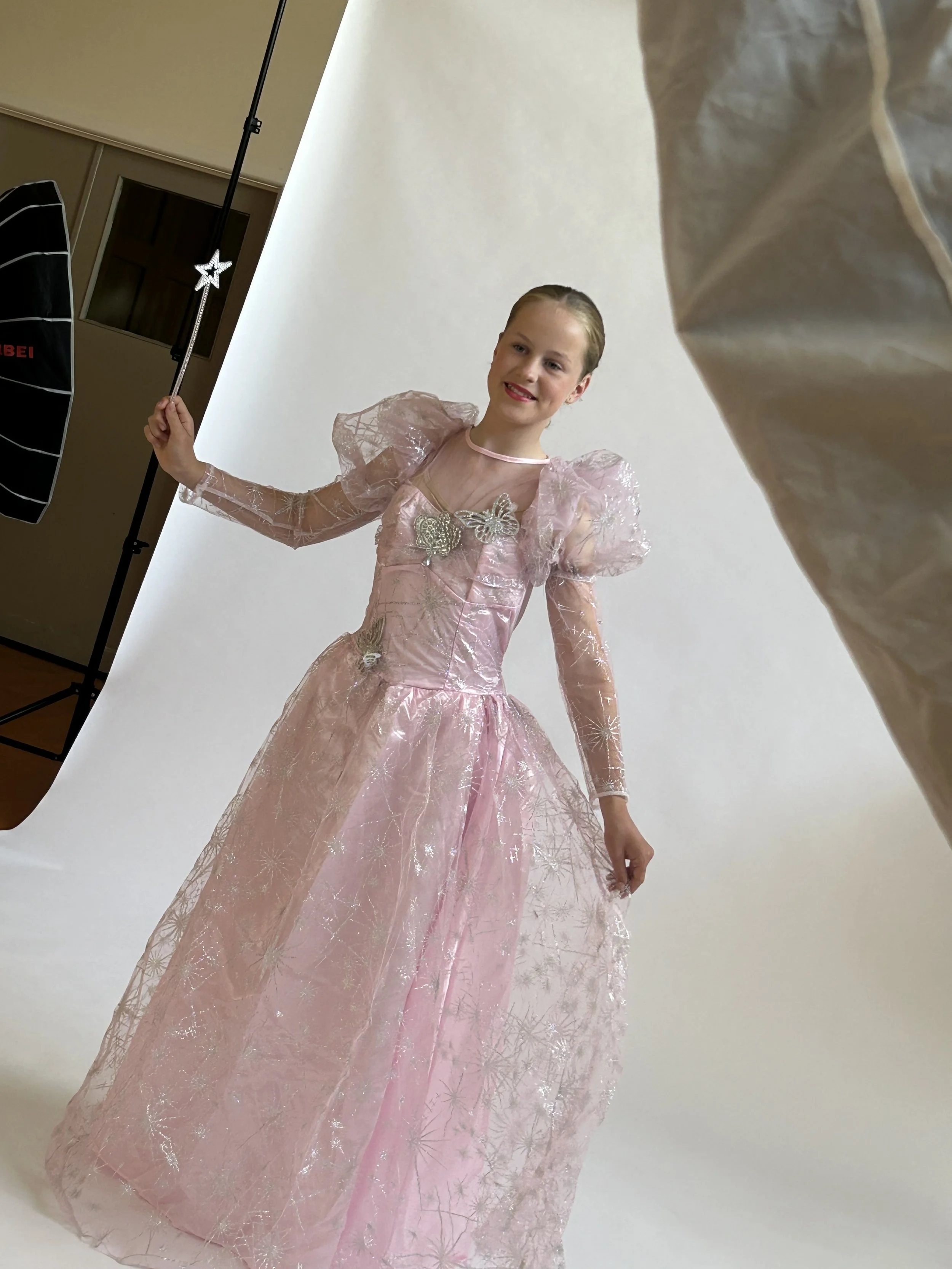 A young girl in a pink fairy-tale princess dress posing in a photography studio with a white backdrop, holding a star-shaped wand, smiling.