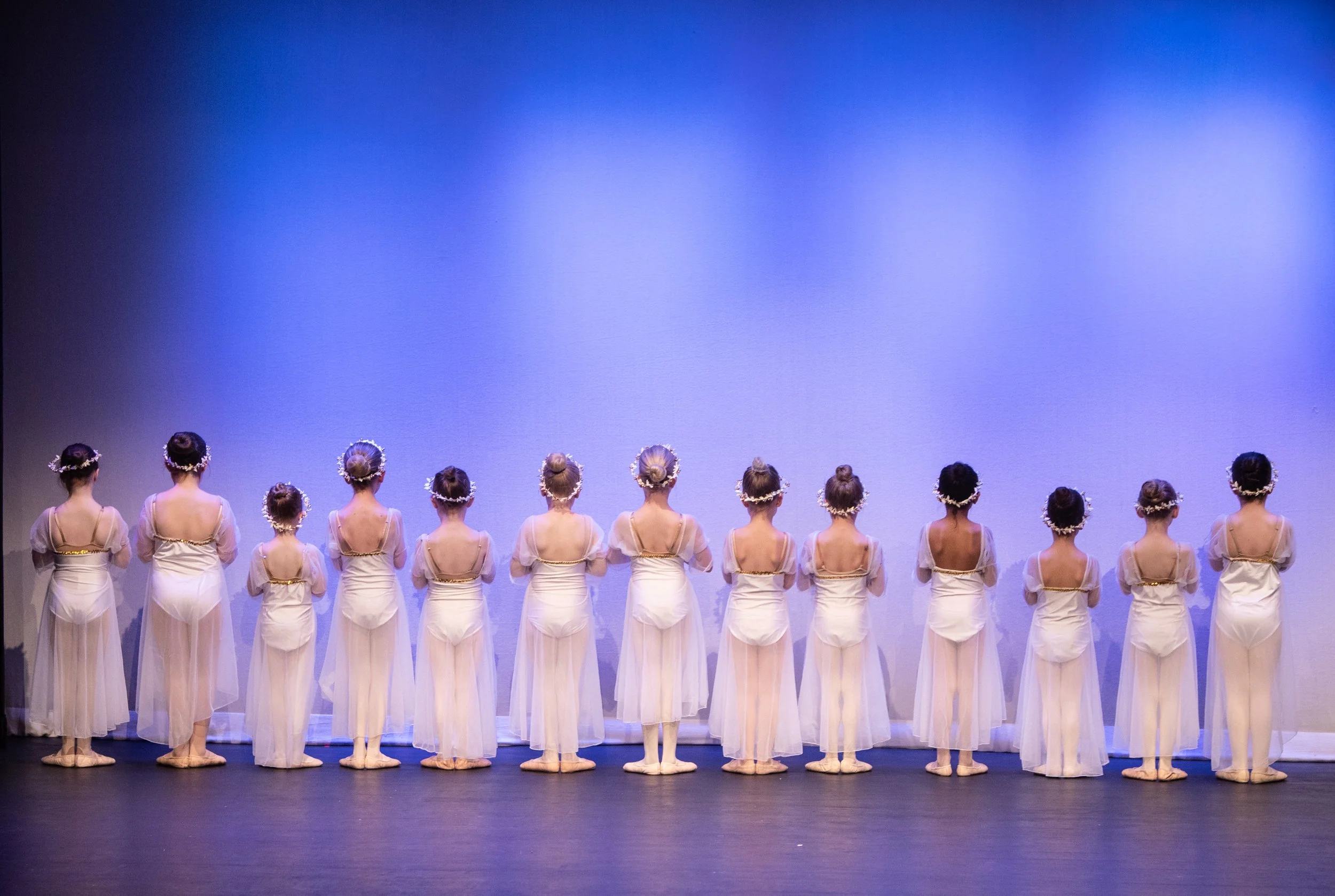 A line of young ballet dancers in white costumes, standing with their backs to the camera on stage, with a blue background.