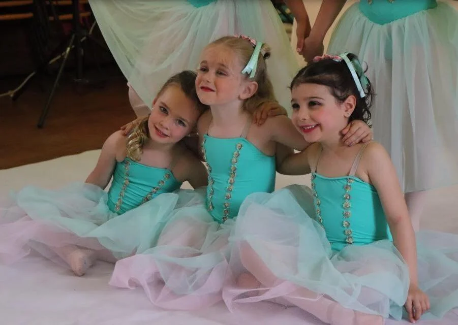 Three young girls dressed as ballerinas in matching turquoise tutus and tops, sitting on the floor at a dance recital or performance.