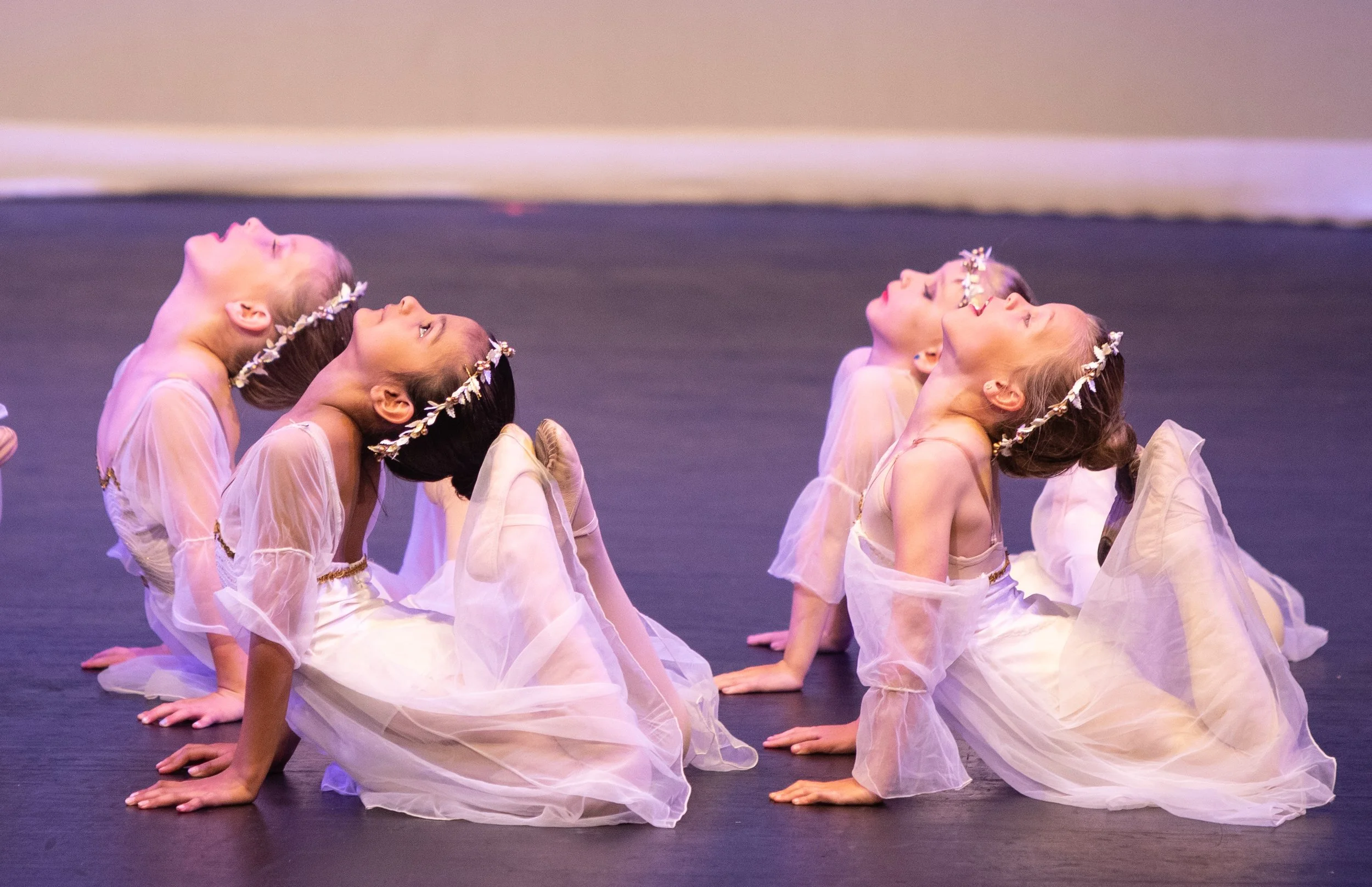 Young girls in ballet costumes performing a backbend pose on stage, wearing flower crowns and cream-colored dresses with flowing skirts.