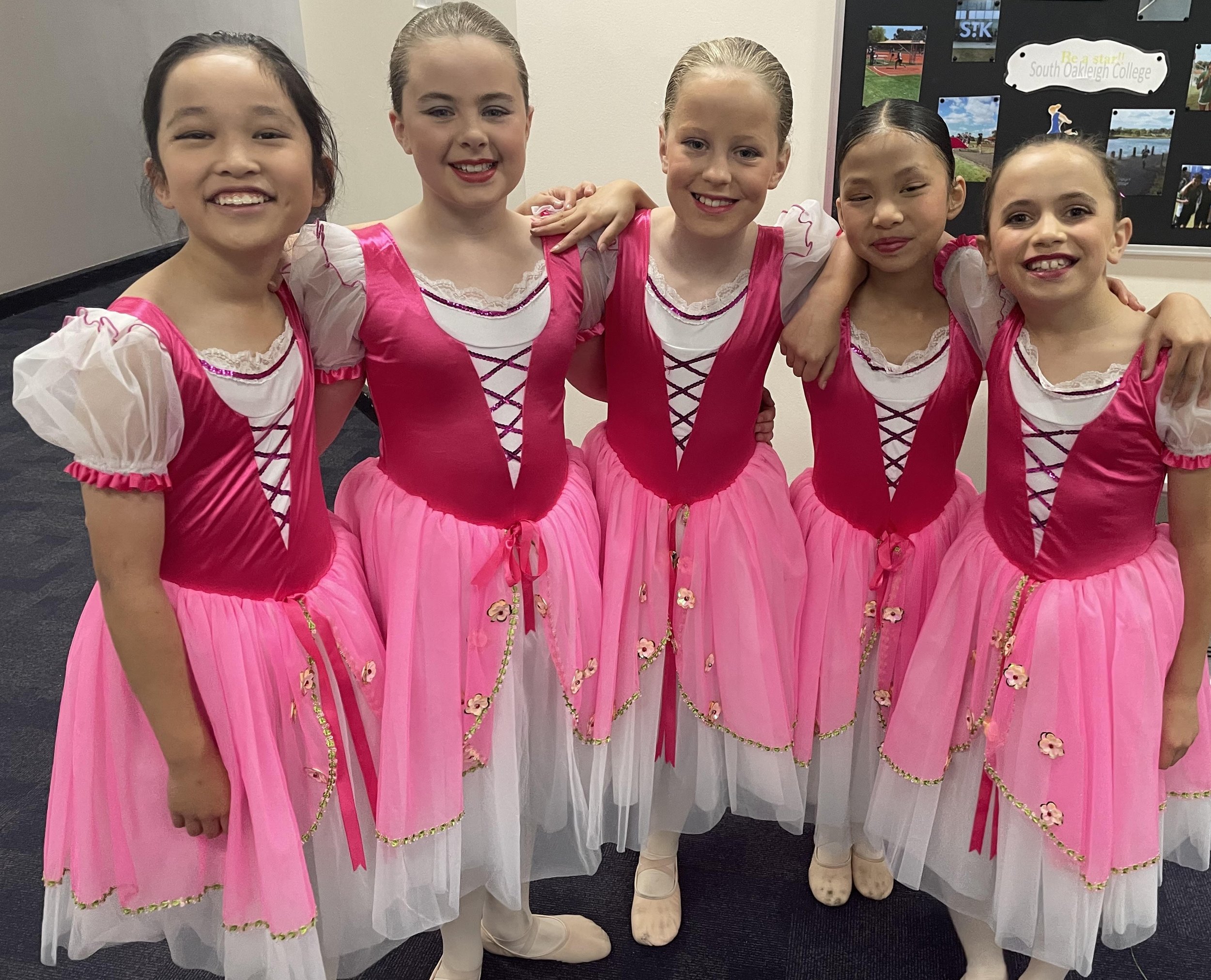 Five young girls dressed in pink and white ballet costumes, standing side by side with their arms around each other, smiling at the camera.