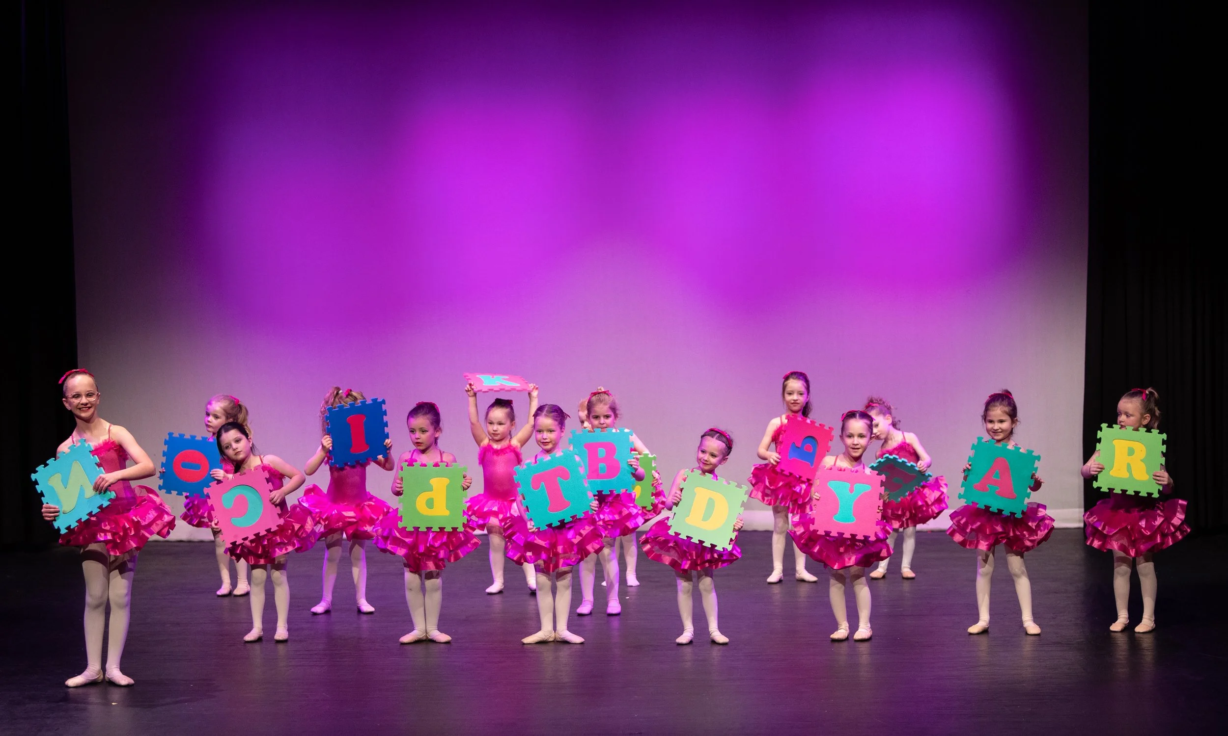 Young girls in pink ballet costumes holding colorful foam puzzle pieces with letters on a stage during a dance performance.