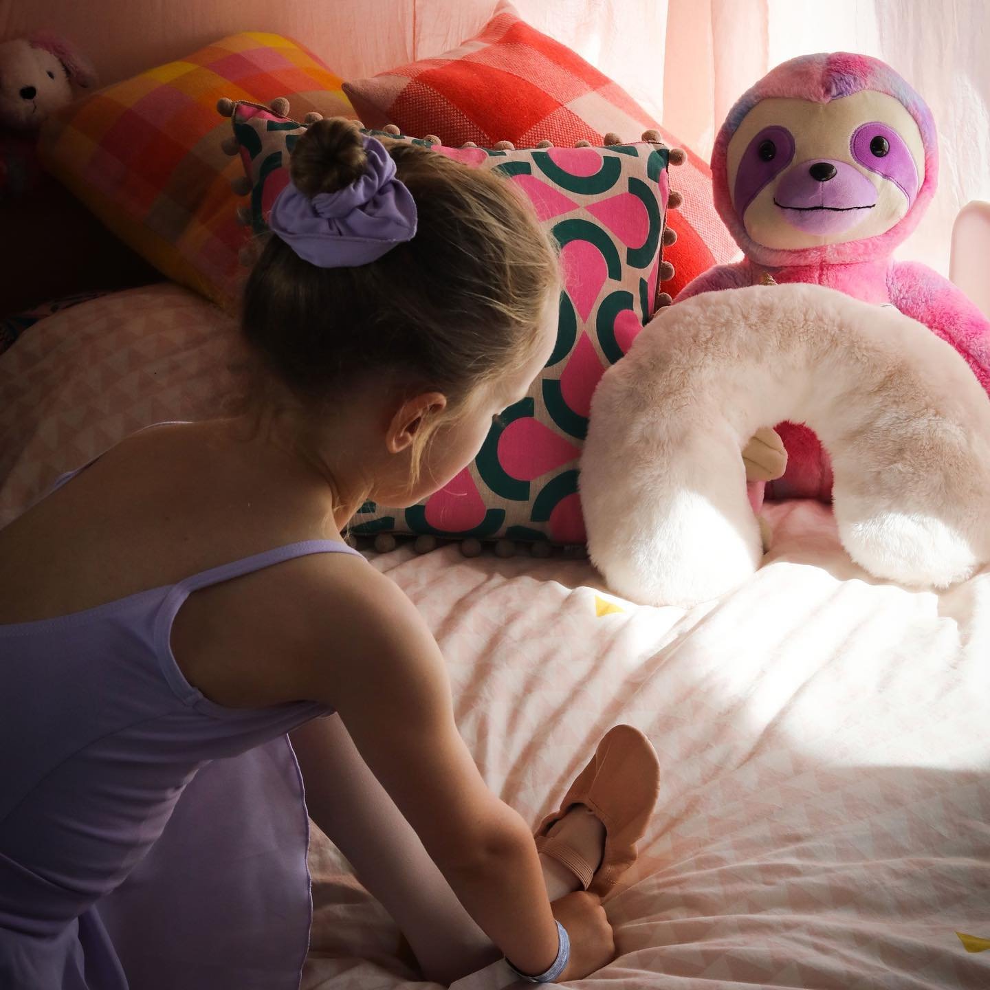 A young girl is sitting on a bed, tying her shoe near colorful pillows and plush toys, including a pink sloth and a beige ring-shaped plush.