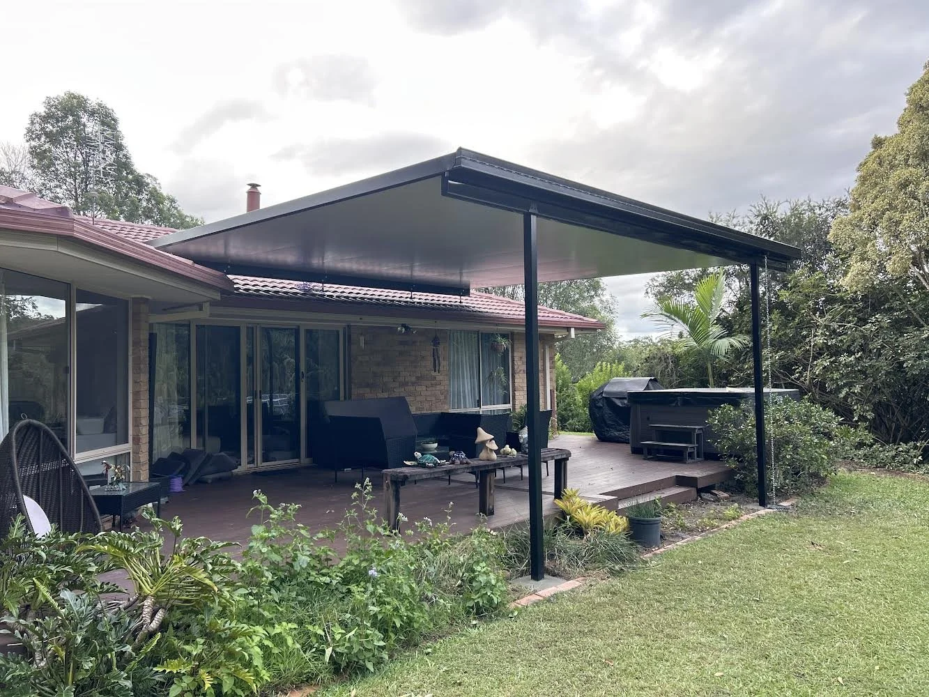 Backyard patio with outdoor furniture, a hot tub covered with a black cover, potted plants, and surrounding greenery in a suburban backyard.