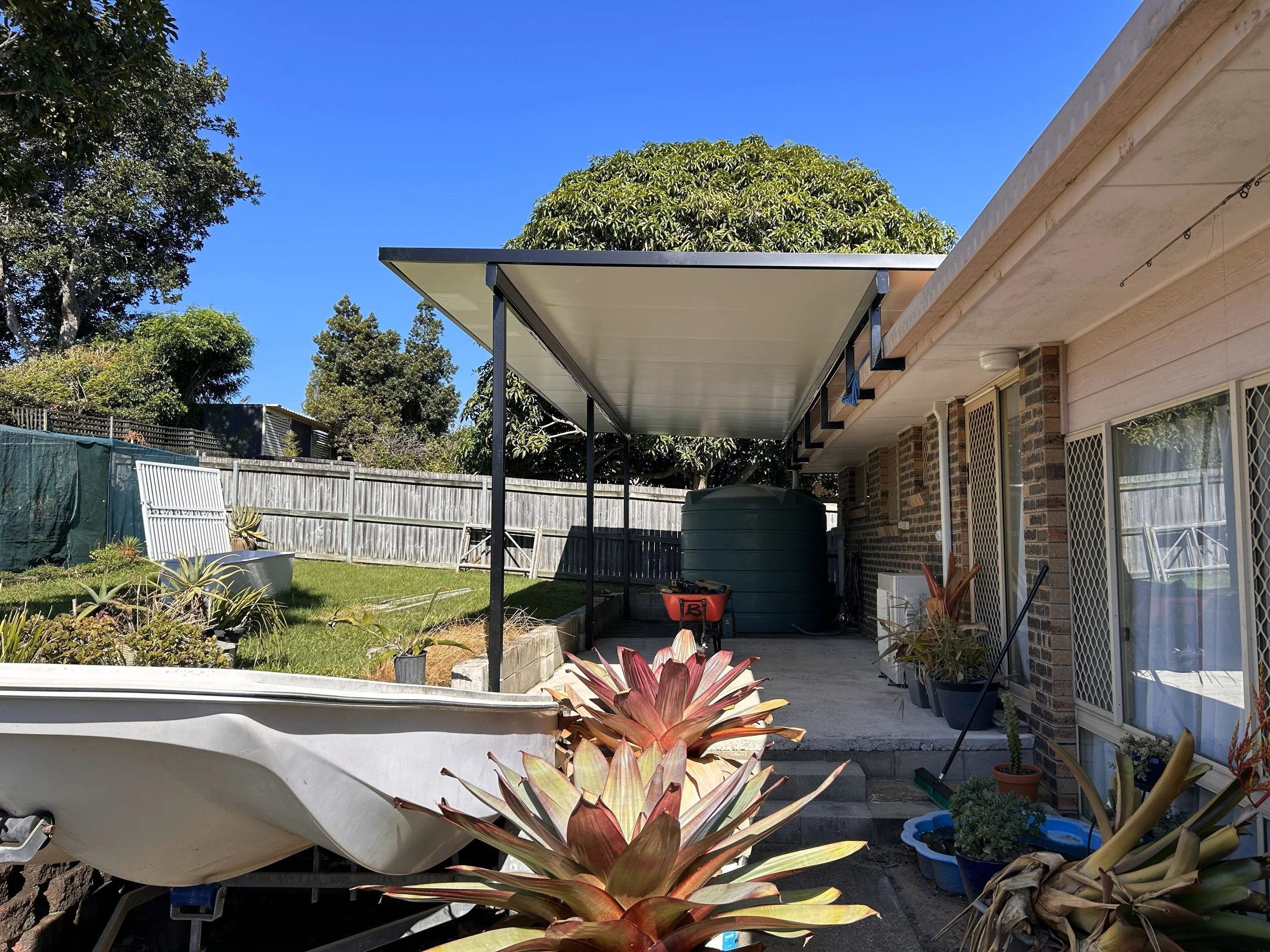 Backyard patio with plants, garden bed, water tank, and covered seating area under a blue sky.