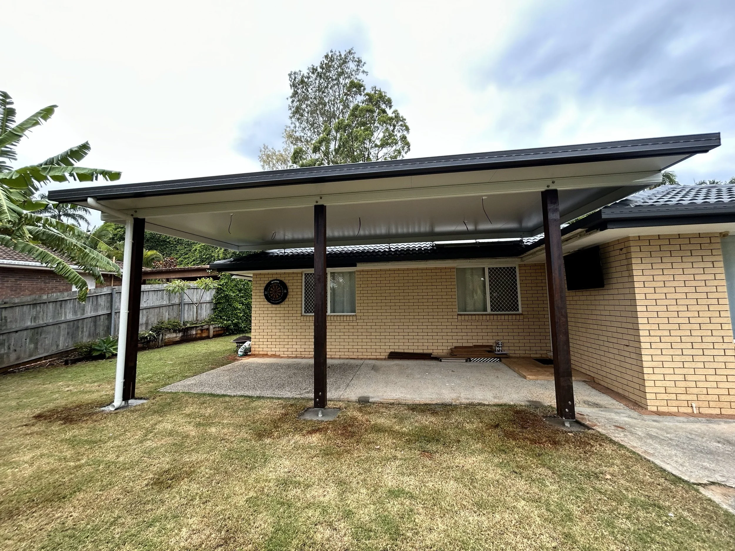Backyard of a house with a brick exterior, a covered patio area with wooden support beams, a dartboard on the wall, and a flat screen TV in the corner.