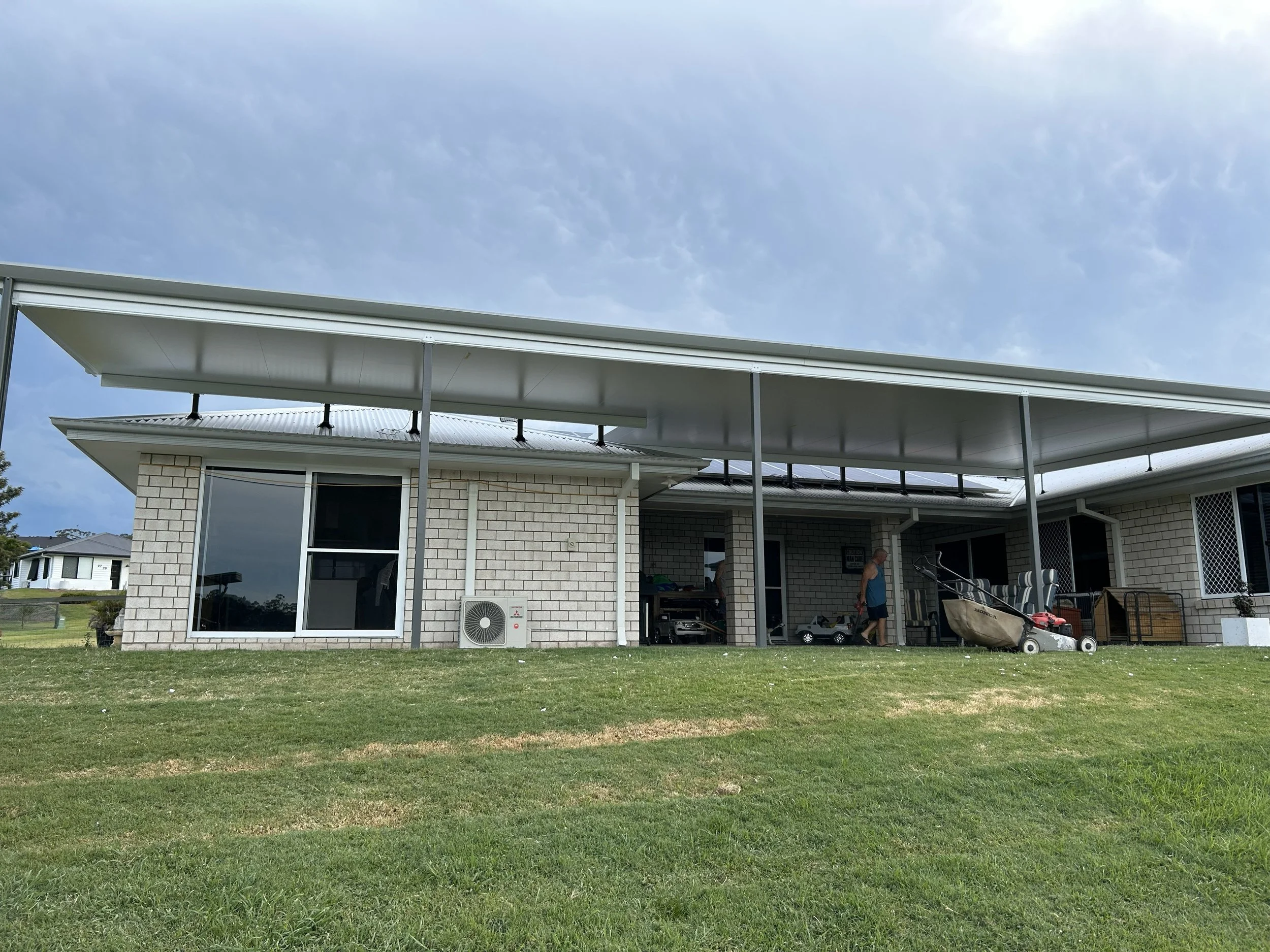 Single-story house with white brick exterior, large glass sliding door, and a covered patio area with a lawn in the foreground. A person is walking near the patio with various lawn equipment and furniture visible.