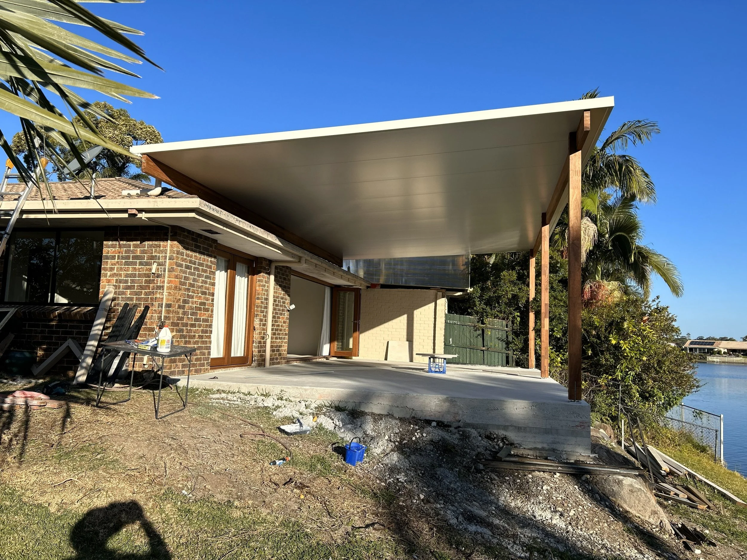 Construction site on a patio with a concrete foundation, brick house, large white roof extension supported by wooden posts, and a view of water and trees in the background.