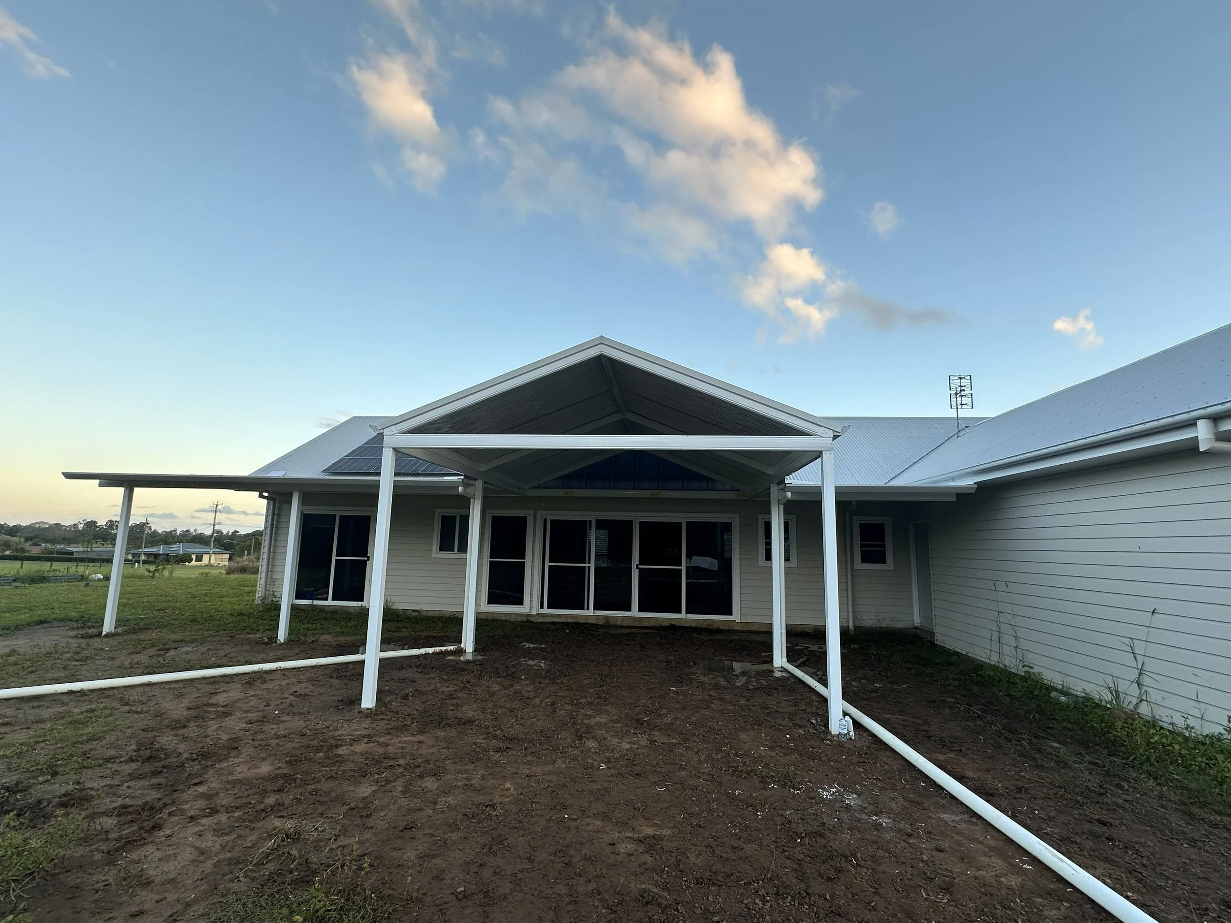 Back of a house under construction with white siding and a metal roof, with plumbing pipes on the ground, on a clear day with a blue sky and some clouds.