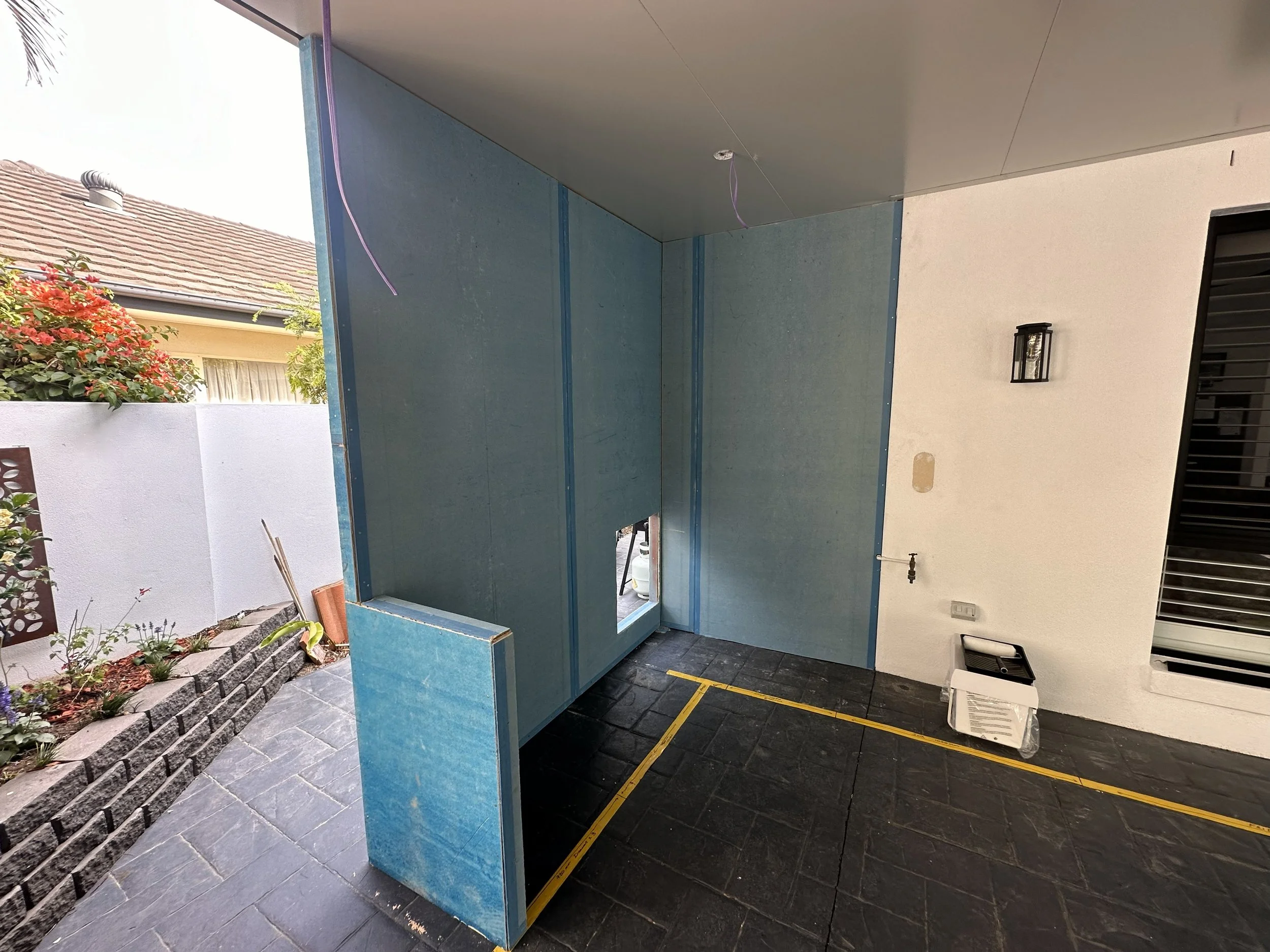 Under construction or renovation on an outdoor patio with cement tiles, with a partially built wall with blue drywall, a small window opening, and construction tools and materials.