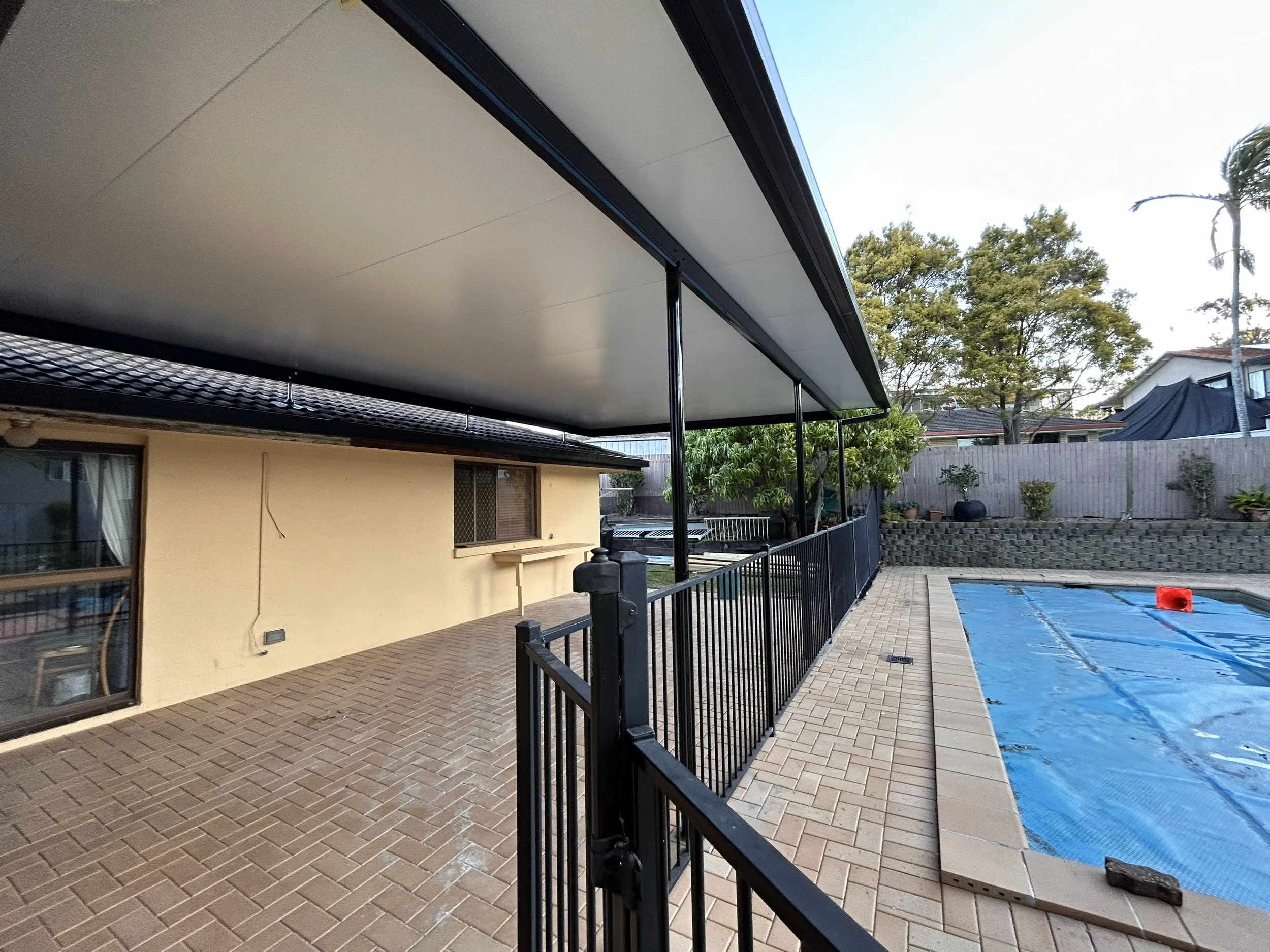 Empty backyard with swimming pool covered by a blue tarp, surrounded by a brick patio and a black metal fence, with trees and neighboring houses in the background.