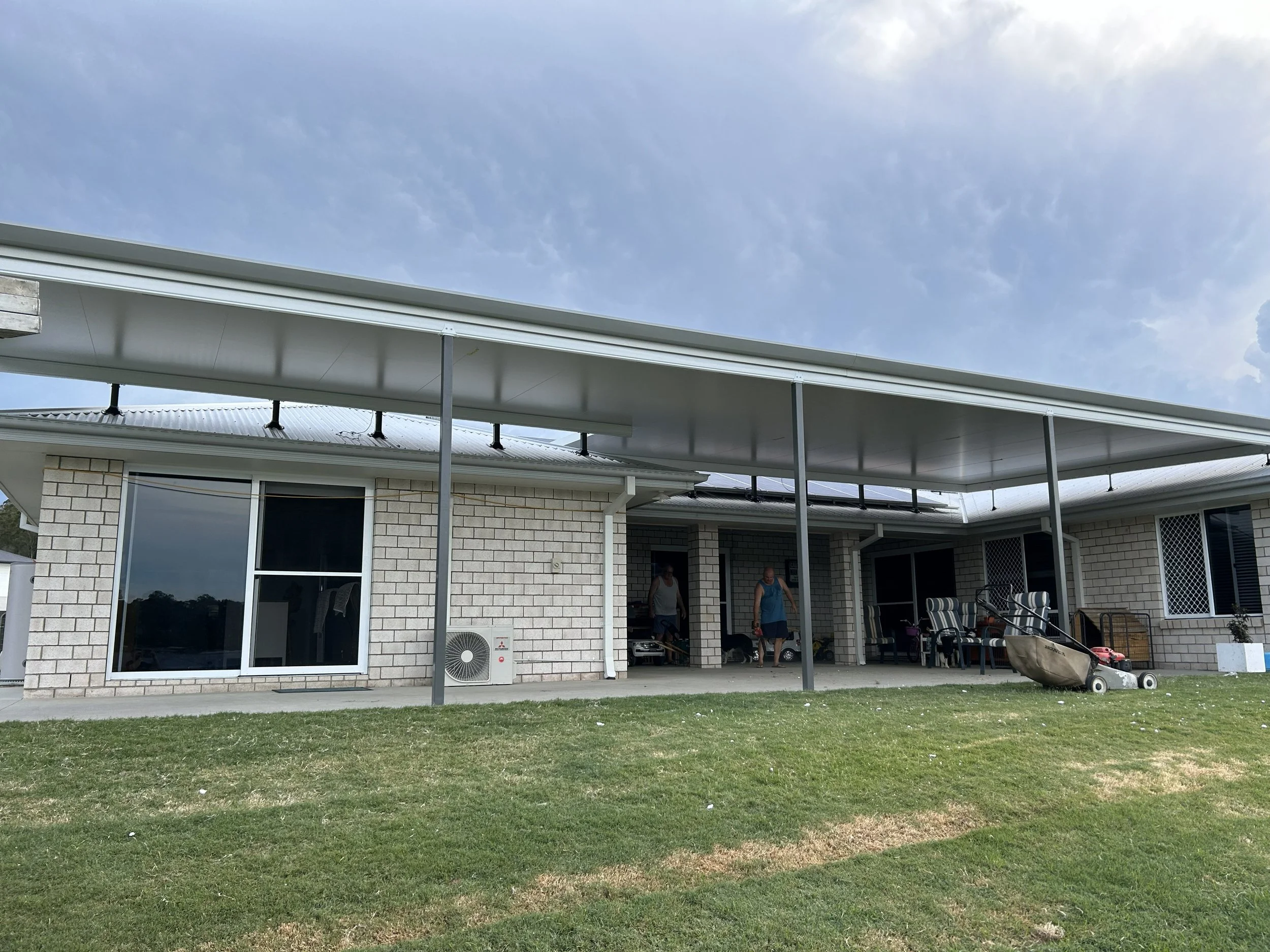 Backyard of a house with a covered patio, lawn, sliding glass doors, and two people walking near the door.