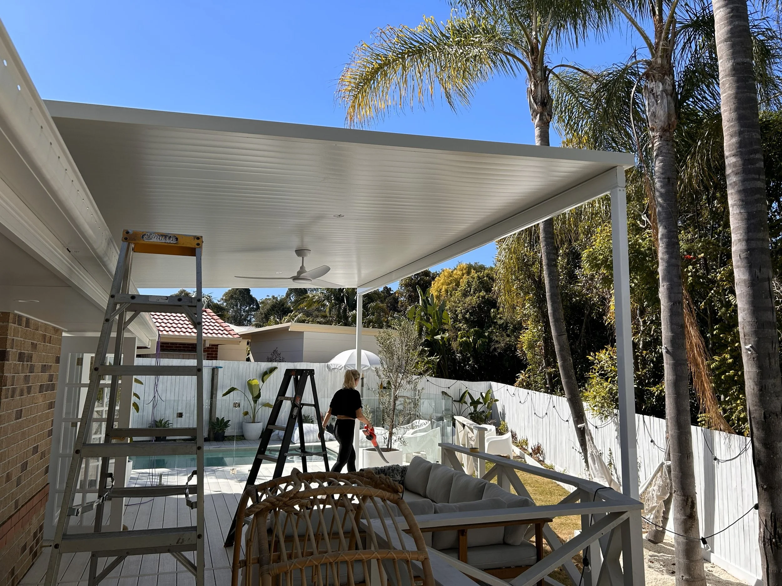 A woman watering plants on a backyard patio with a white ceiling, ceiling fan, glass railing, and tall palm trees, during sunny weather.