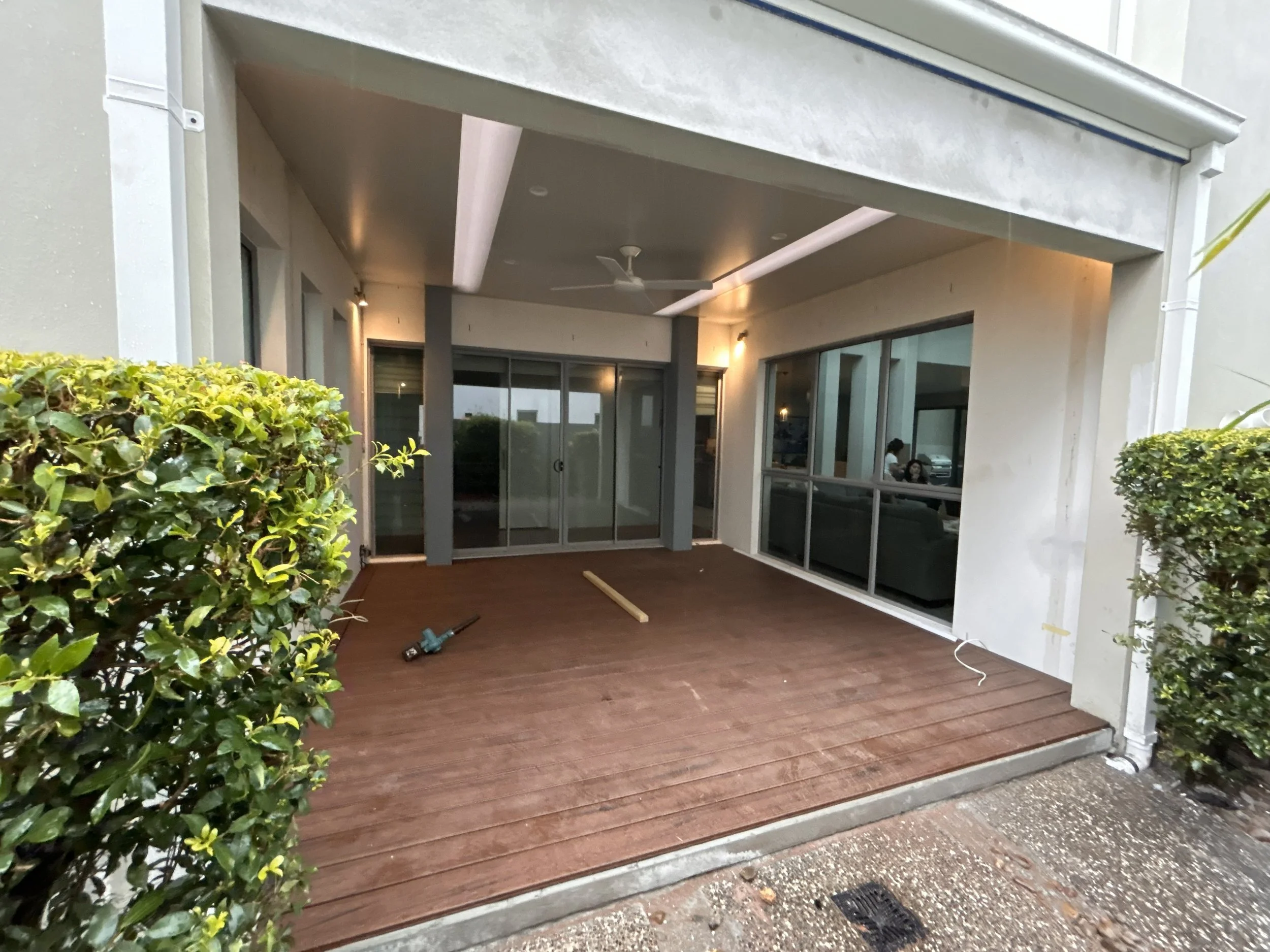 Back porch with wooden decking, glass sliding doors, and patio furniture inside. Two bushes on either side of the porch. Construction tools and a piece of wood lying on the deck.