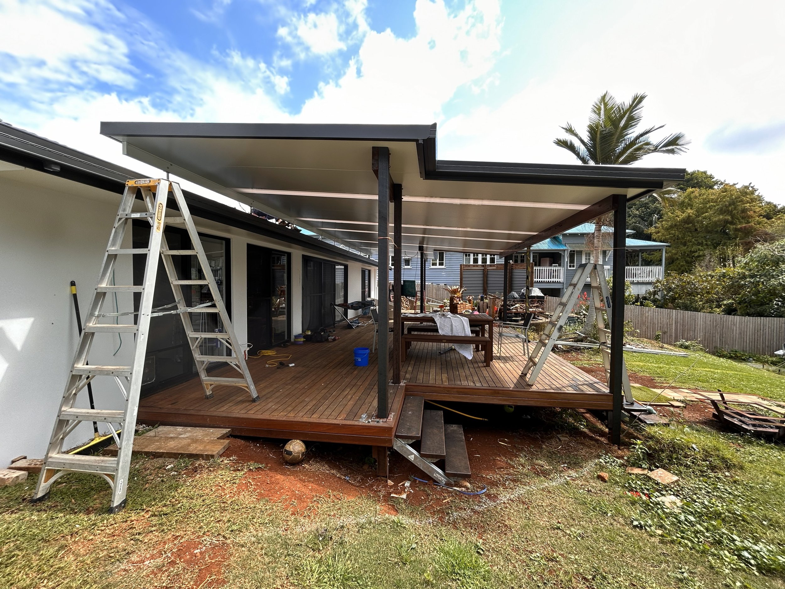 Construction of a covered outdoor deck on the back of a house with ladders, tools, and furniture around.