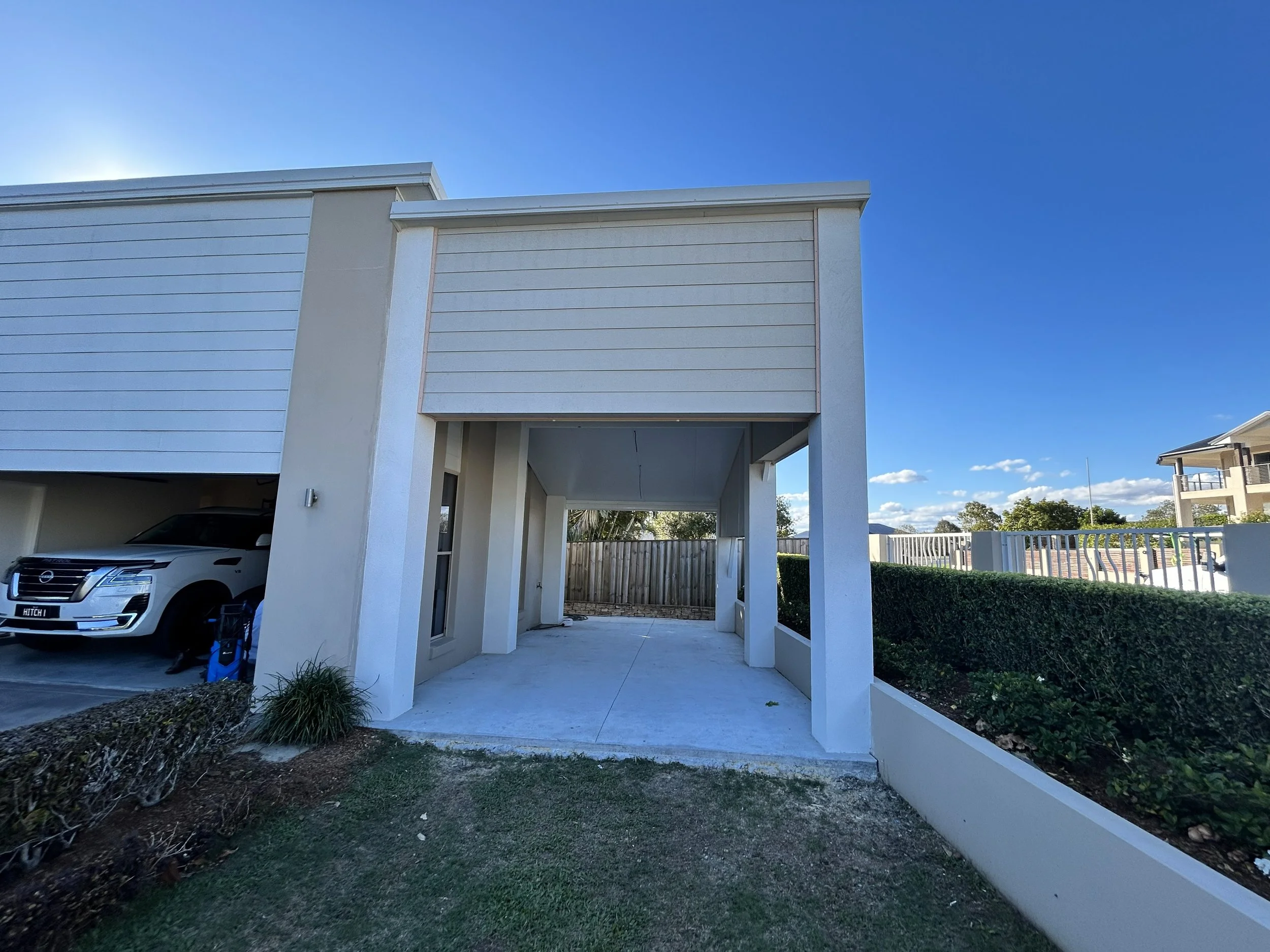 View of a modern residential house with a driveway and carport, white facade, and well-maintained lawn under a clear blue sky.
