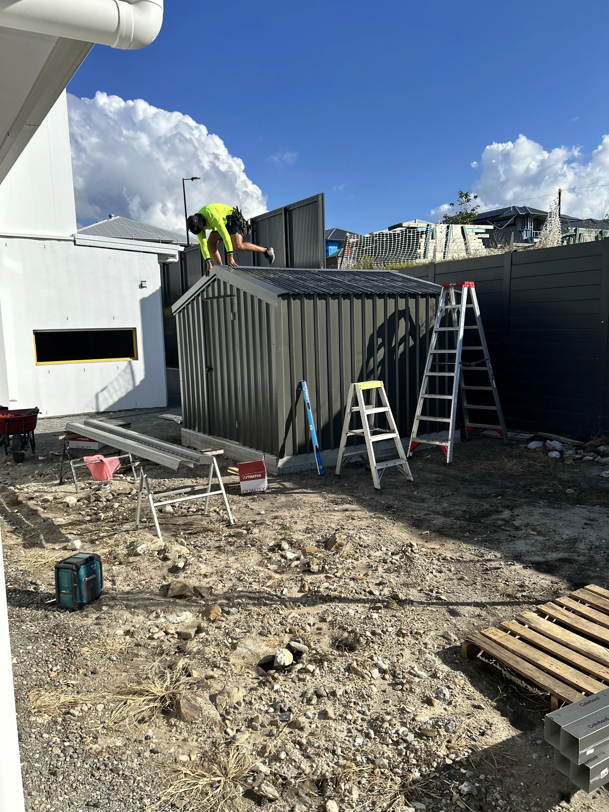 A construction worker in a neon yellow shirt and black helmet is climbing on a metal shed, working on its roof. Various ladders, tools, and construction materials are nearby in an outdoor construction site, with a clear blue sky and clouds overhead.