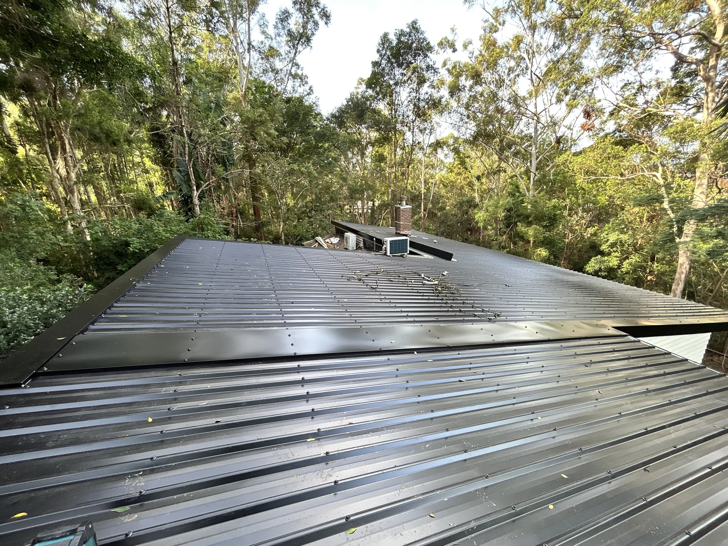 Metal roof with a vent, air conditioning unit, and chimney surrounded by trees.