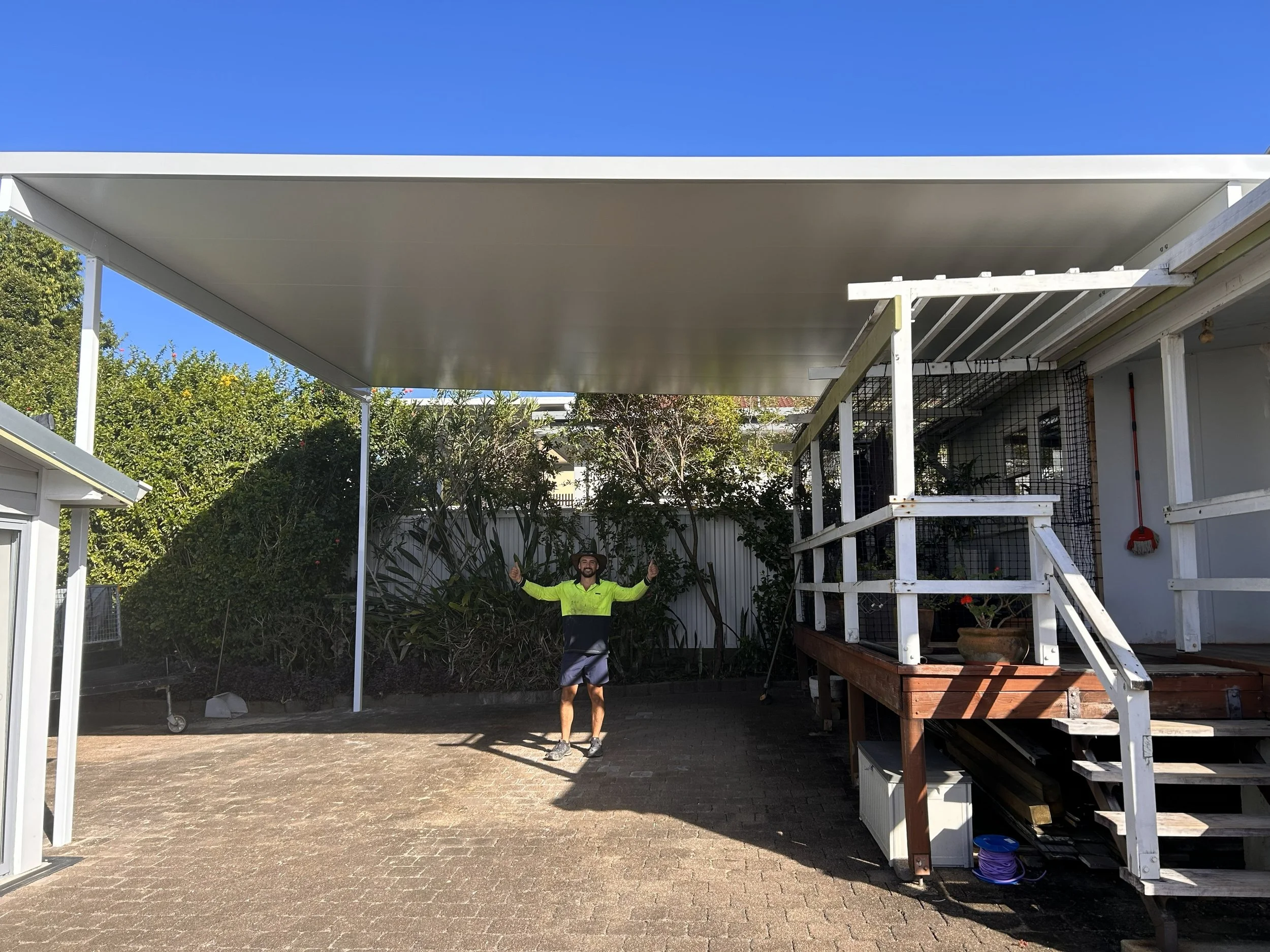 A man standing outdoors on a paved area, smiling with arms raised, wearing a neon yellow and black shirt and shorts, under a large modern white patio roof attached to a house, with a background of trees and a clear blue sky.