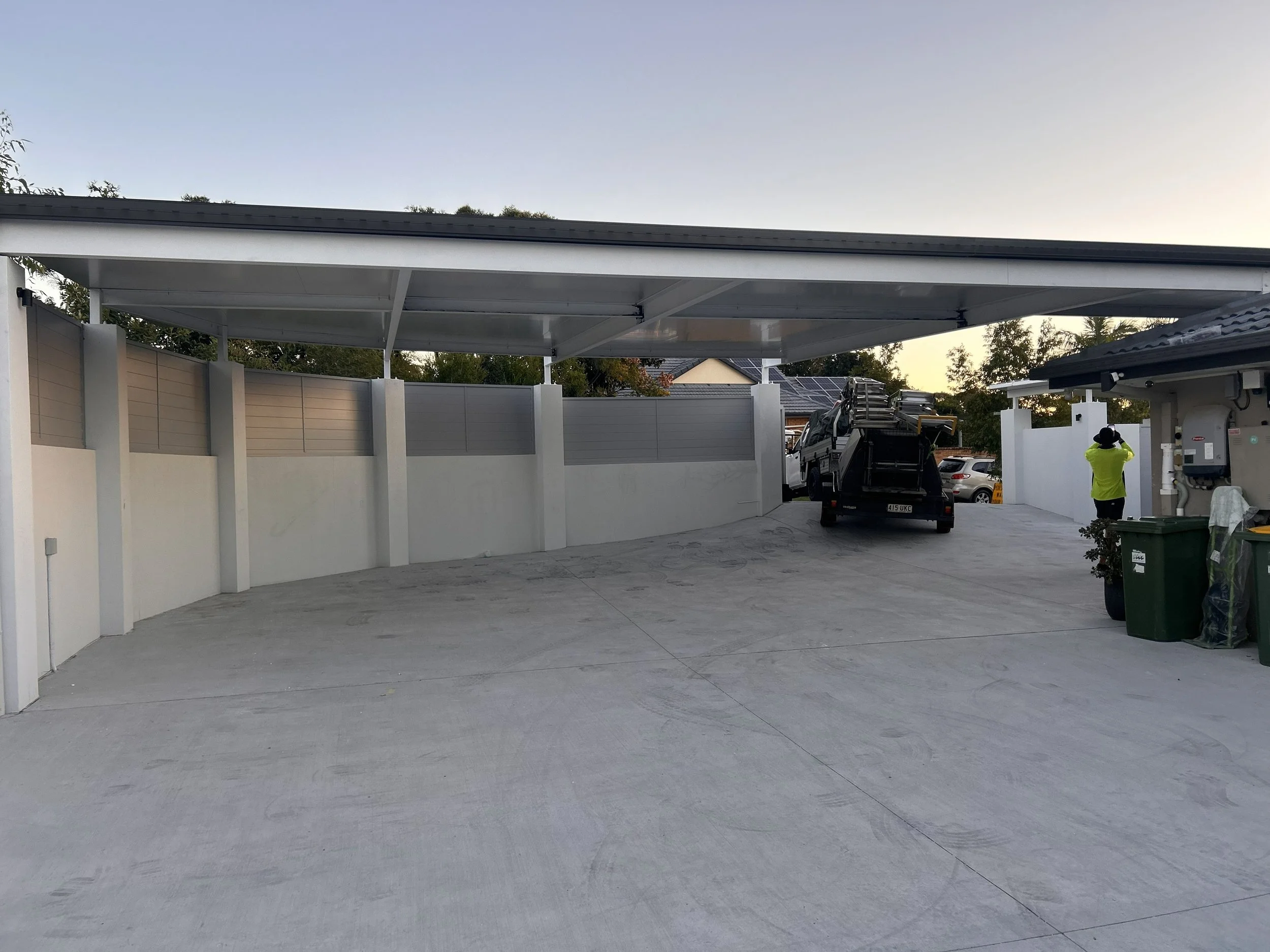 A residential driveway with a covered carport. There are a black pickup truck on a flatbed trailer and a person wearing a yellow shirt standing near garbage bins.