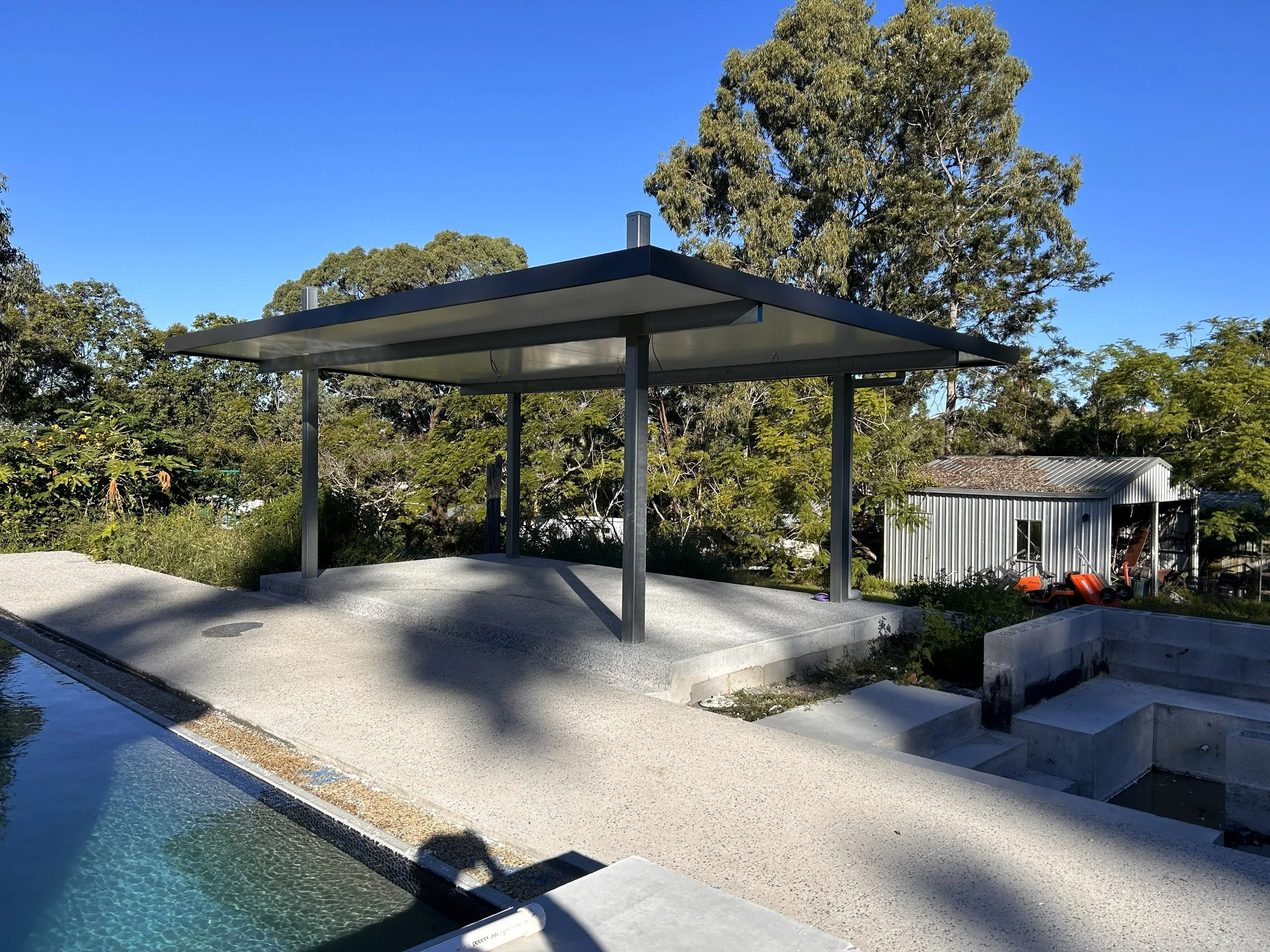 Modern outdoor covered pavilion with a black metal roof supported by several black metal posts, situated over a concrete platform near a swimming pool; background includes trees and a small shed with gardening equipment.