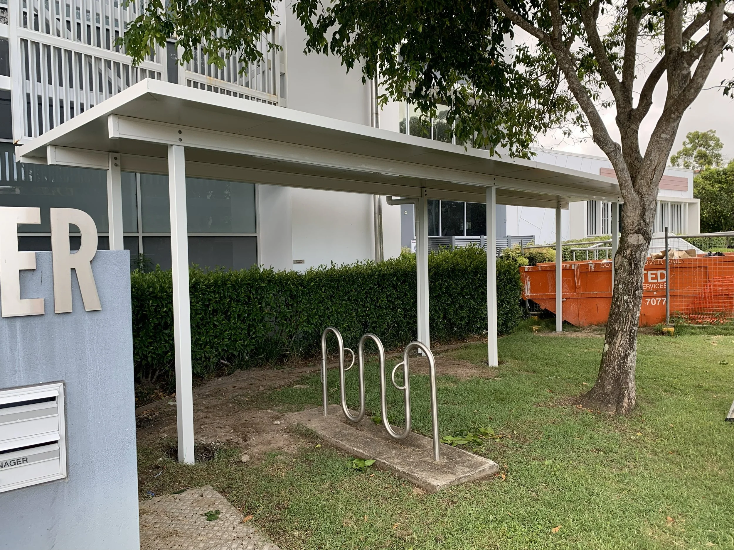 Bike rack with two metal loops under a white canopy next to a tree and a hedge, in front of a building with large glass windows.