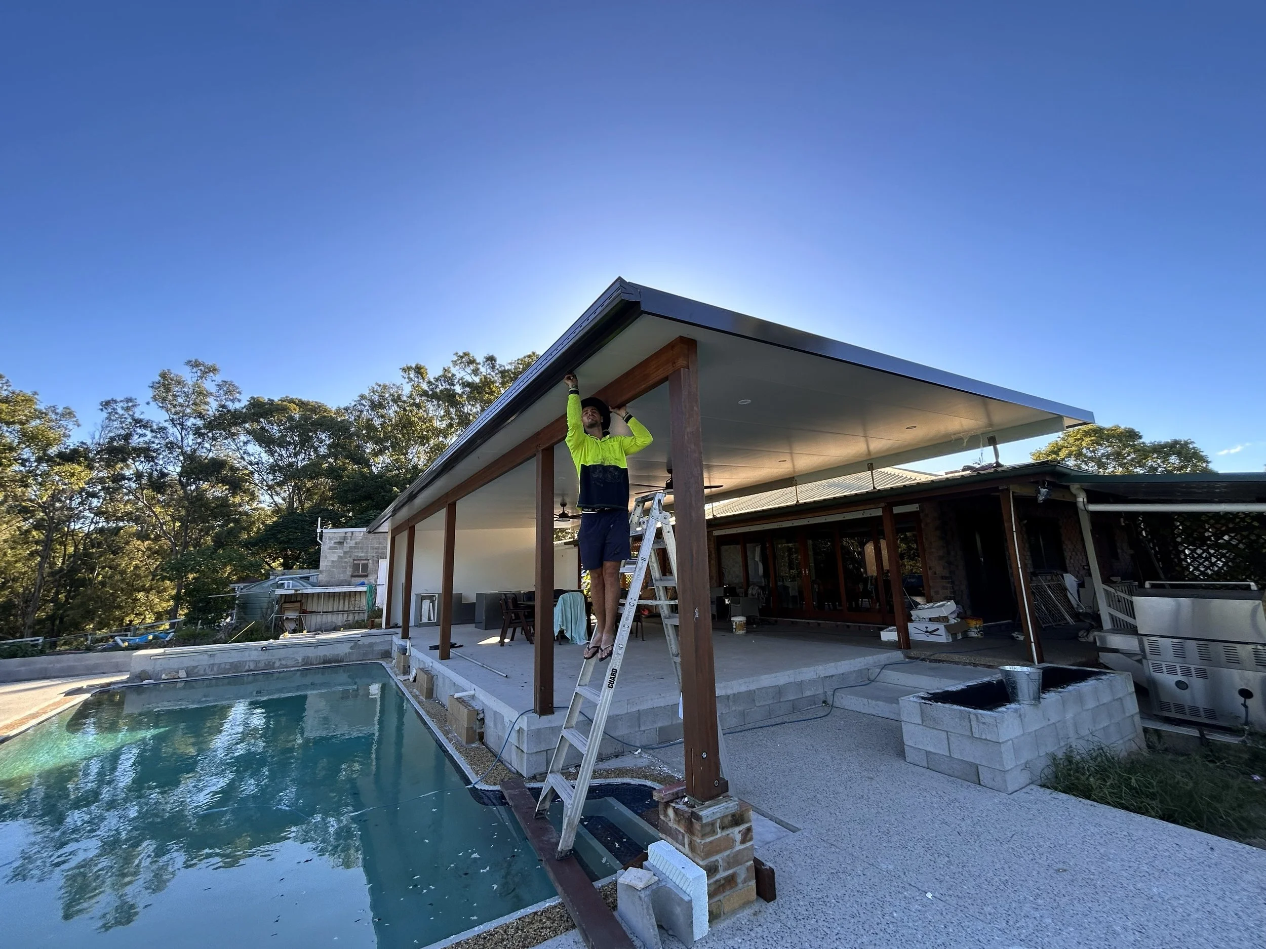 A man wearing a neon yellow and black long-sleeve shirt and shorts standing on a ladder, working on the roof of a modern house with a pool in the backyard during daylight.